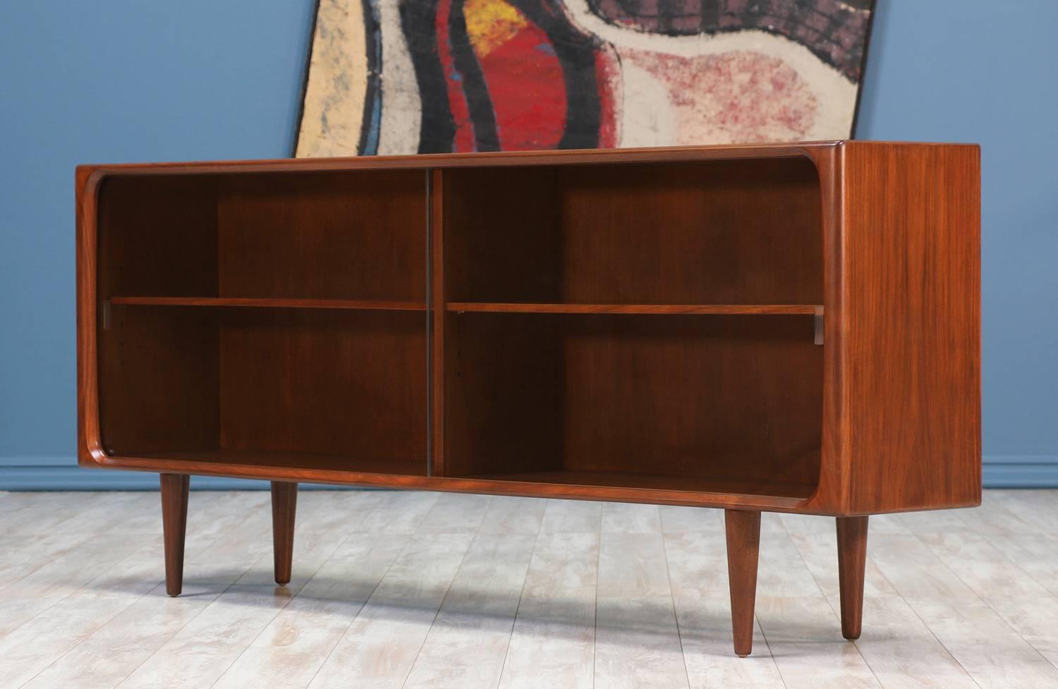 Danish Modern Bookcase designed and manufactured in Denmark circa 1960's. Beautifully crafted with a walnut wood frame and tapered legs, this piece is narrow in design with removable shelves and maintains its original glass doors showing minor