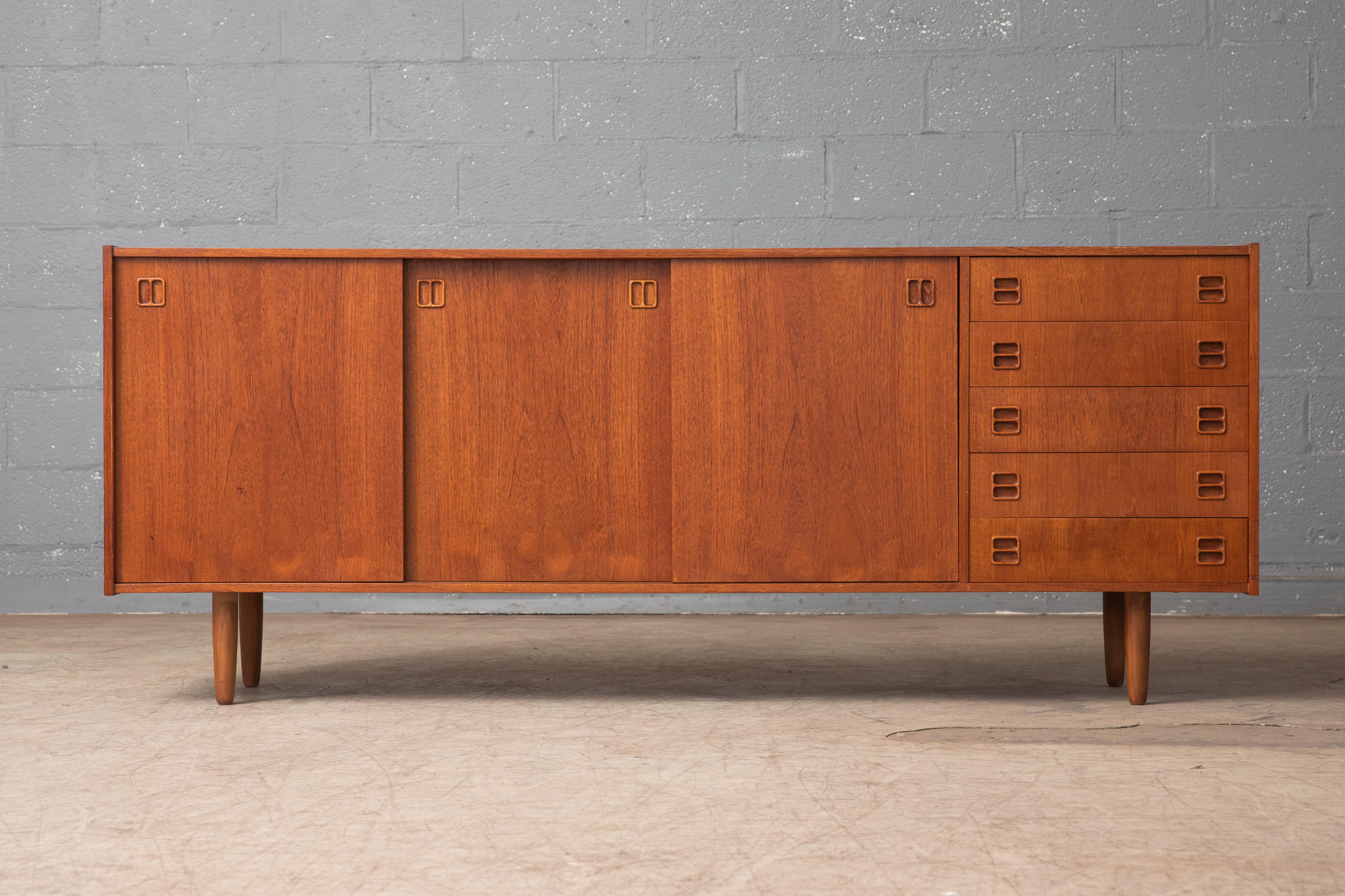 Beautiful Danish midcentury low sideboard in teak made 1960s in the style of Kai Kristiansen. Nice off-center drawer section flanked by compartments with adjustable shelves behind nice bookmatched sliding doors with carved pulls. Very nice color and