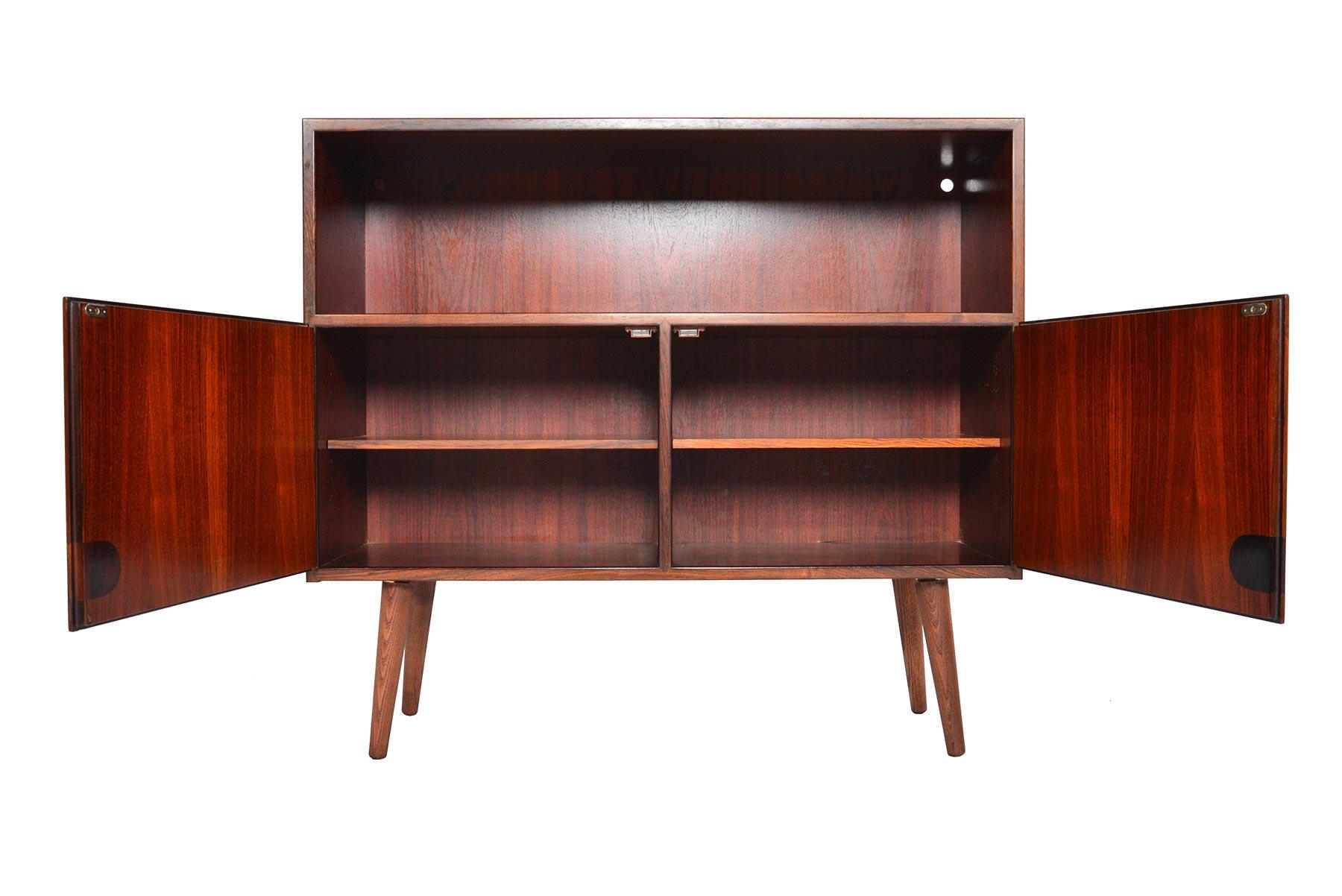This small Danish modern rosewood bookcase is a perfect storage solution for any modern home. This piece features a narrow profile with an open cubby and two lower doors concealing two bays with adjustable shelving. Piece stands on canted spindle