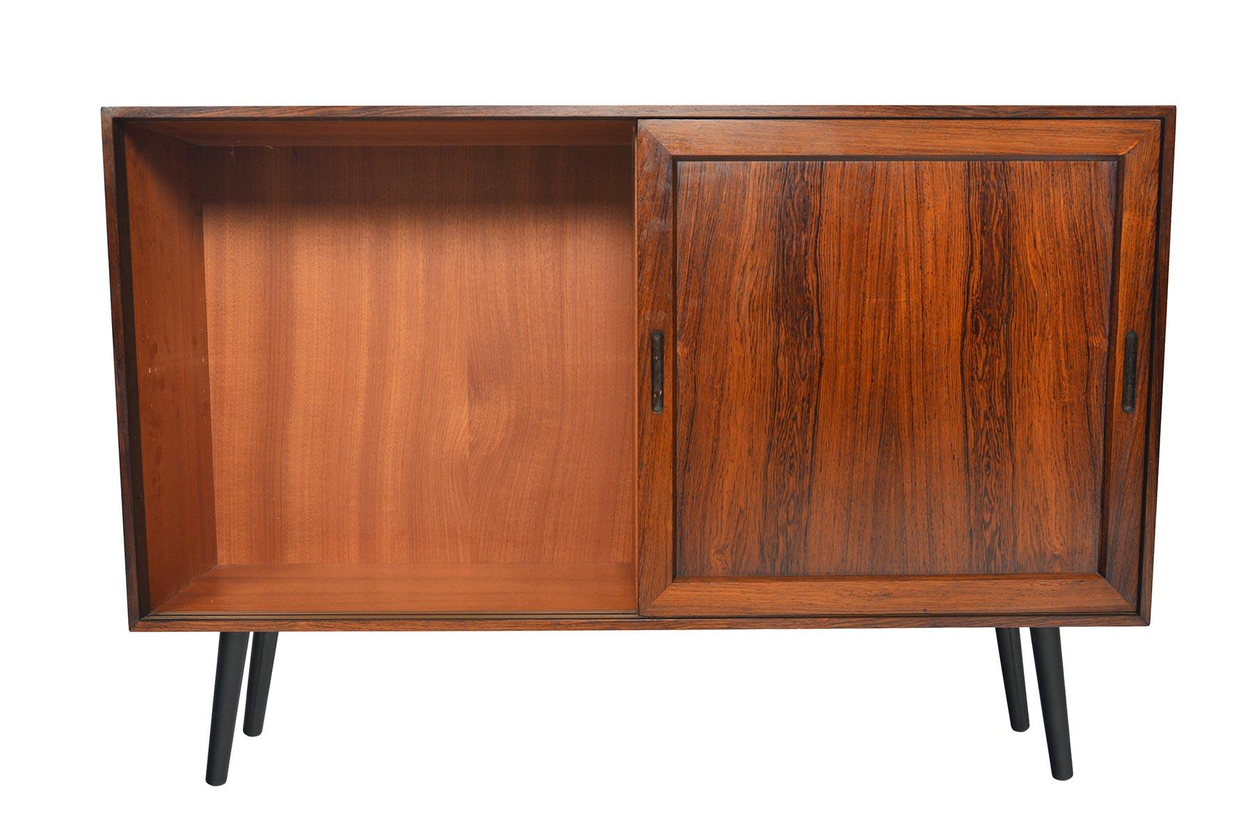 This small Danish modern rosewood credenza with doors is a perfect storage solution for any modern home. This piece features a narrow profile with two bays and adjustable shelving for each side. Piece stands on canted spindle legs. In excellent