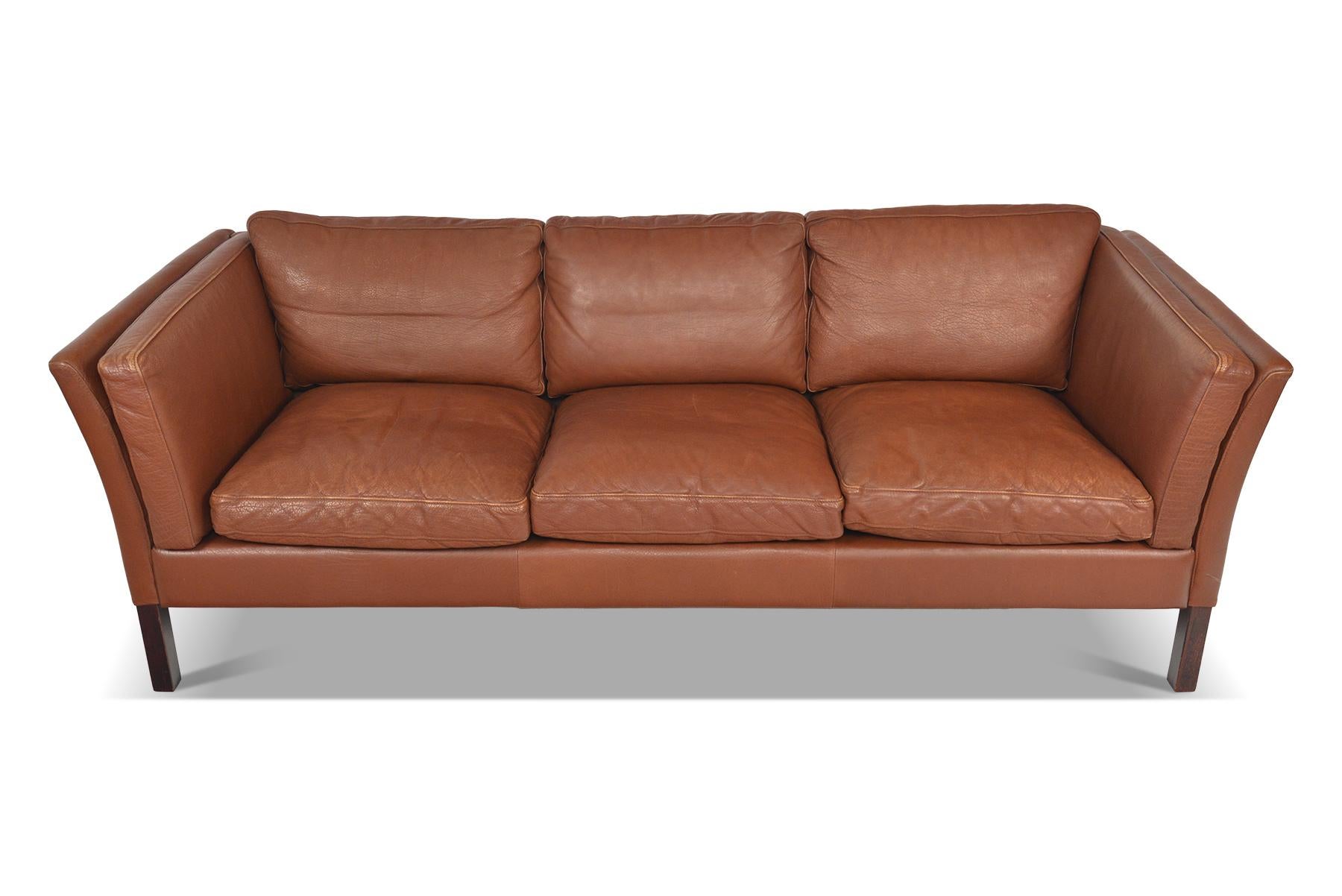 This Danish modern sofa is covered in original patinated rust toned leather. The simple bowed arm design offers an elegant profile. Eight cushions line the interior for a comfortable sitting experience. The sofa stands on a tapered stained beech