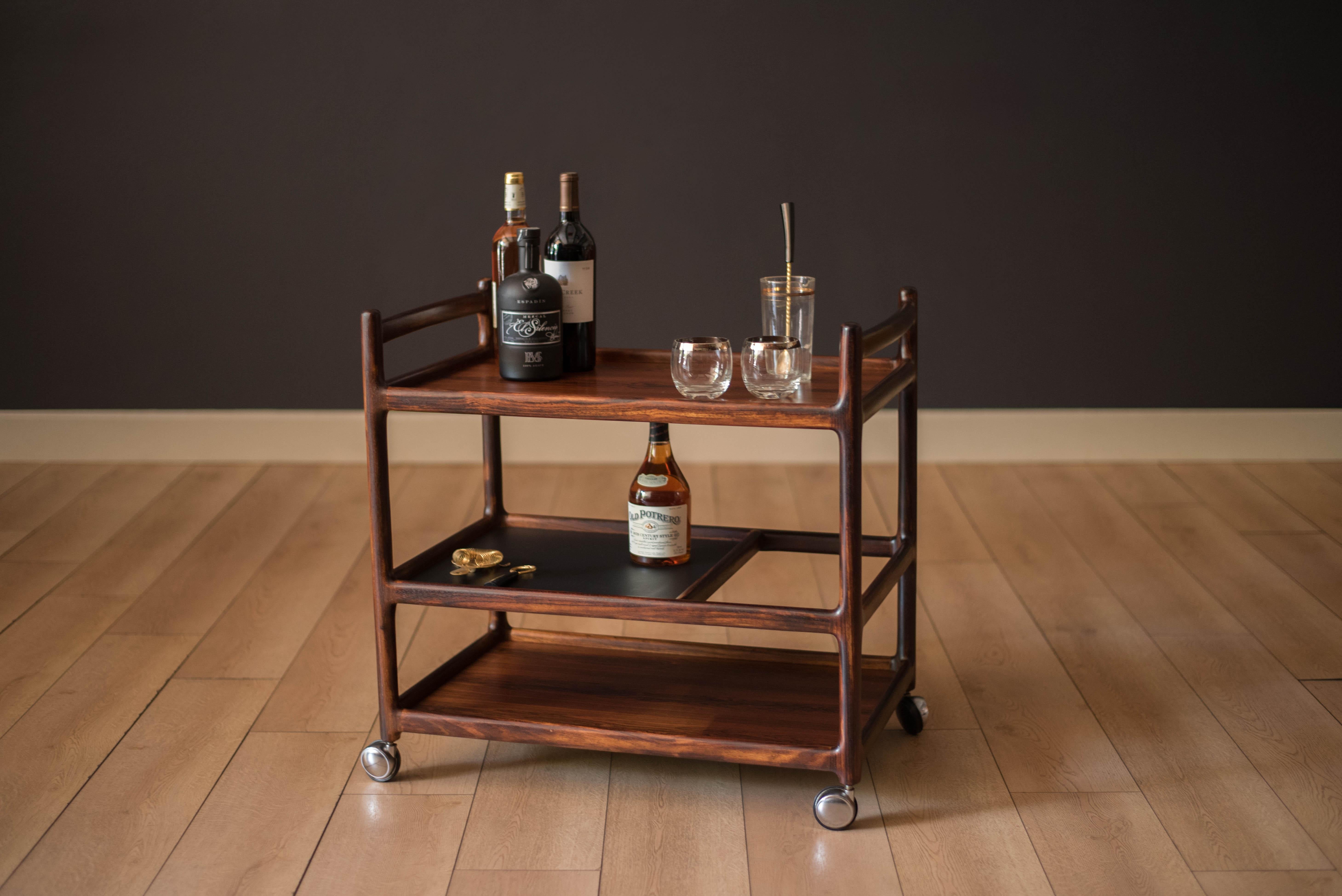 Mid-Century Modern bar cart trolley designed by Johannes Andersen for CFC Silkeborg circa 1960s, Denmark. Features three tiers of shelving storage perfect for tall bottles and glassware. Sculpturally crafted in solid rosewood and includes an easy to