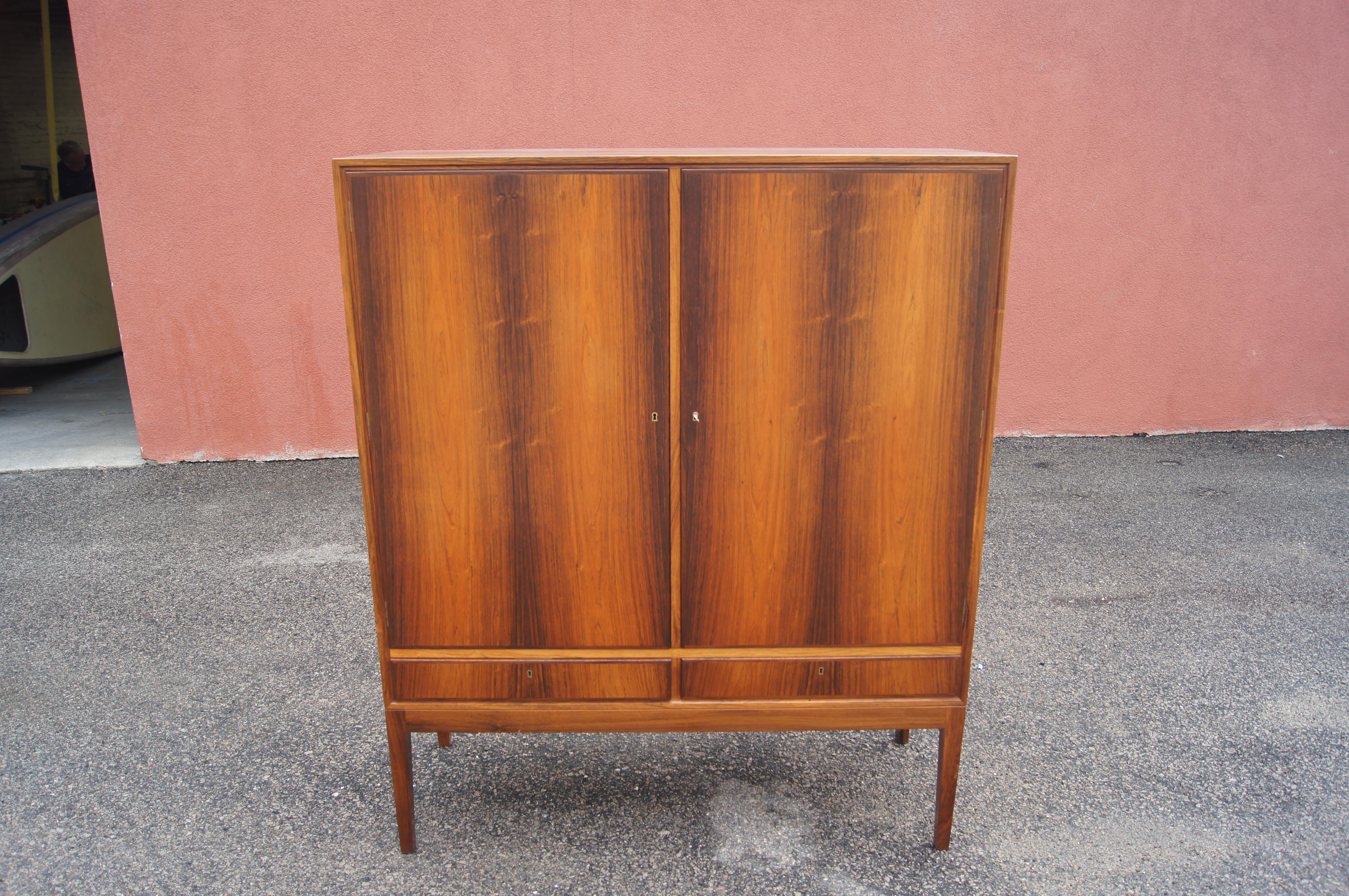 This striking Danish modern cabinet features a tall rosewood case perched on slender legs. Behind the doors (locking with the original key) are two sets of four adjustable shelves; three on the right have tray fronts. At bottom are two drawers.