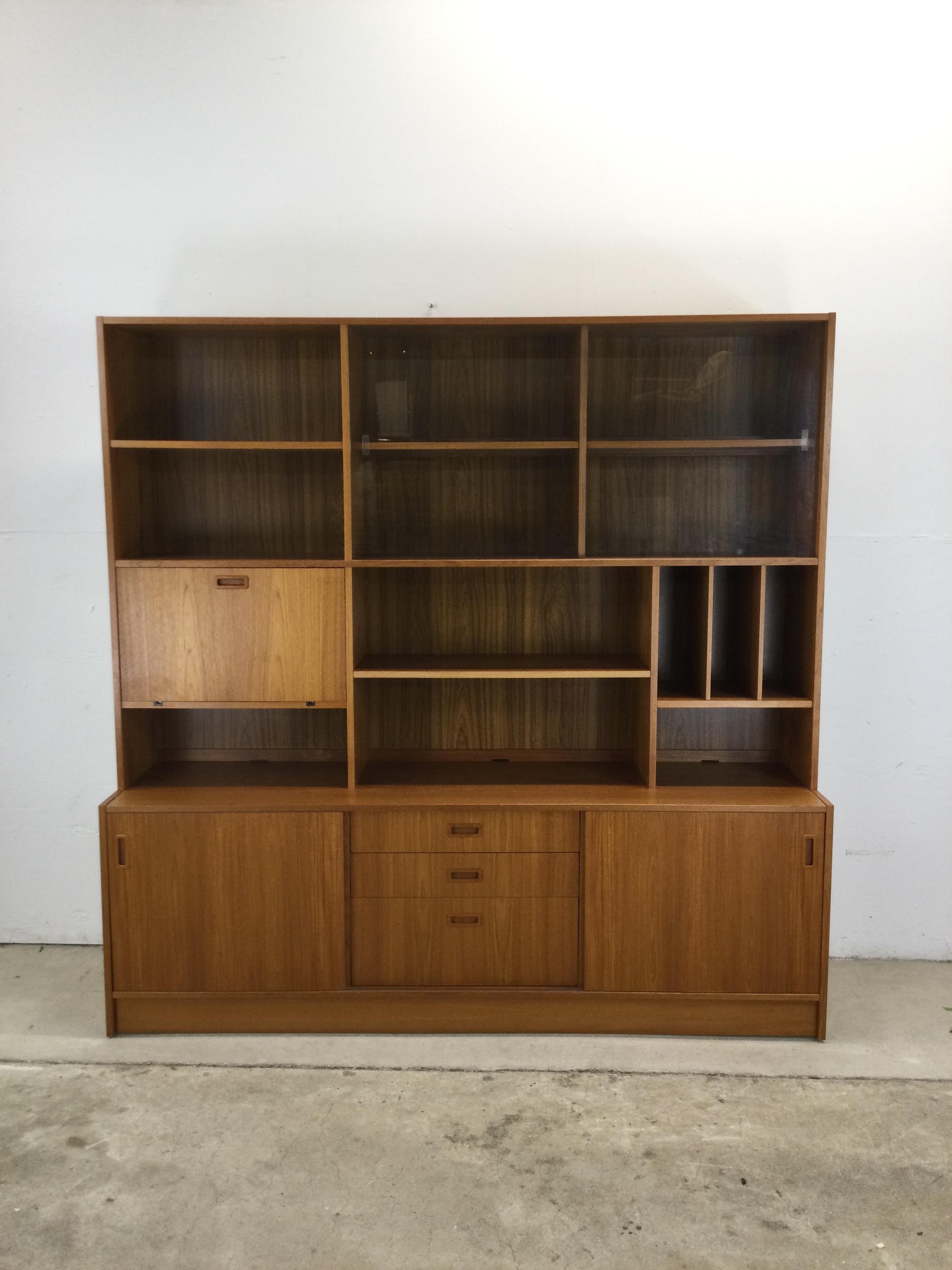 This Danish modern shelving unit features pressed wood construction, teak veneer with original finish, two piece design with adjustable shelving, sliding glass doors, and drop front cabinet up top acco,packed by two sliding doors and three drawers