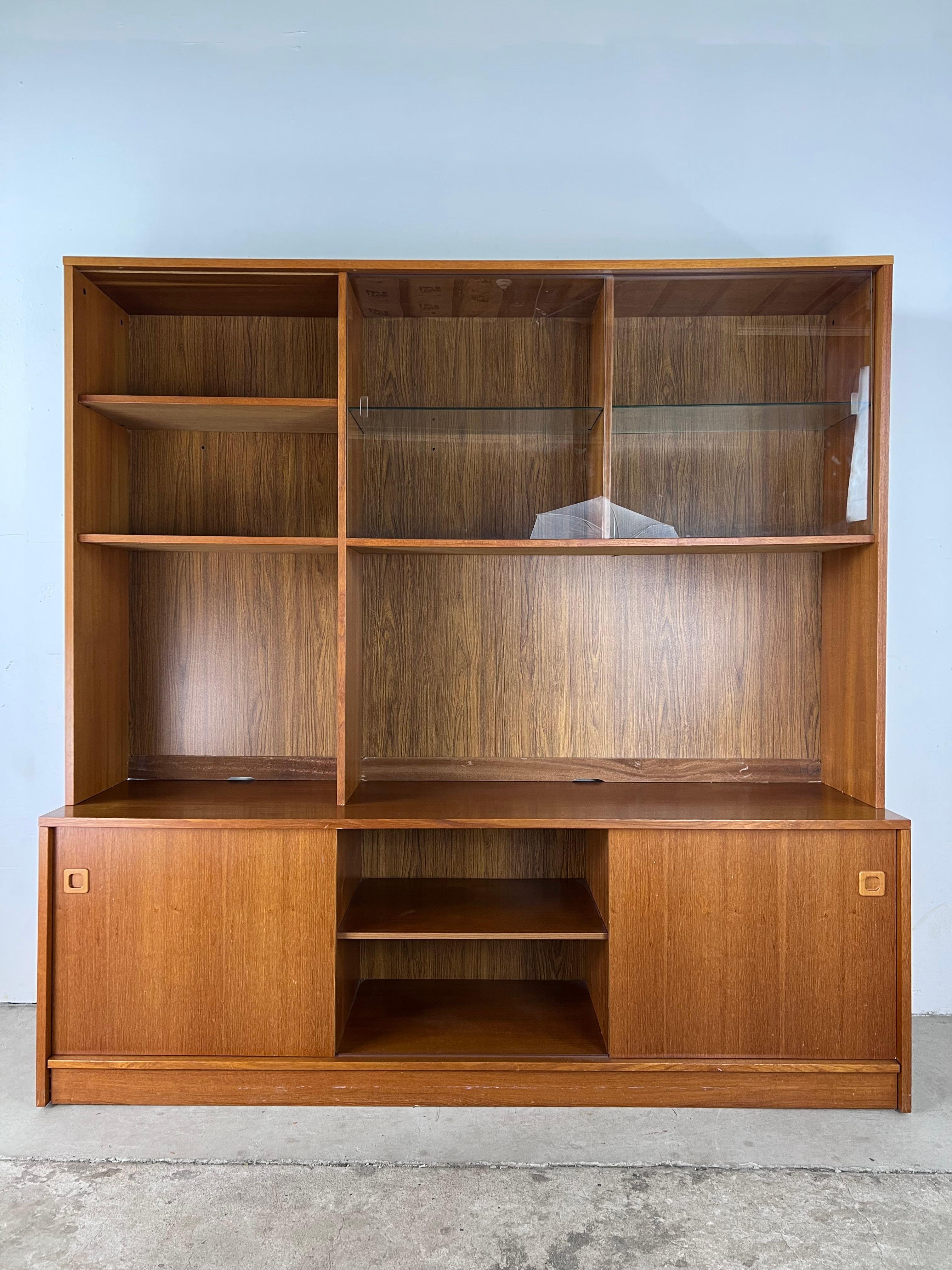 This Danish modern shelving unit features pressed wood construction, teak veneer with original finish, two piece design for easy redecorating, adjustable shelves and sliding glass doors up top, adjustable shelves and sliding wood doors down