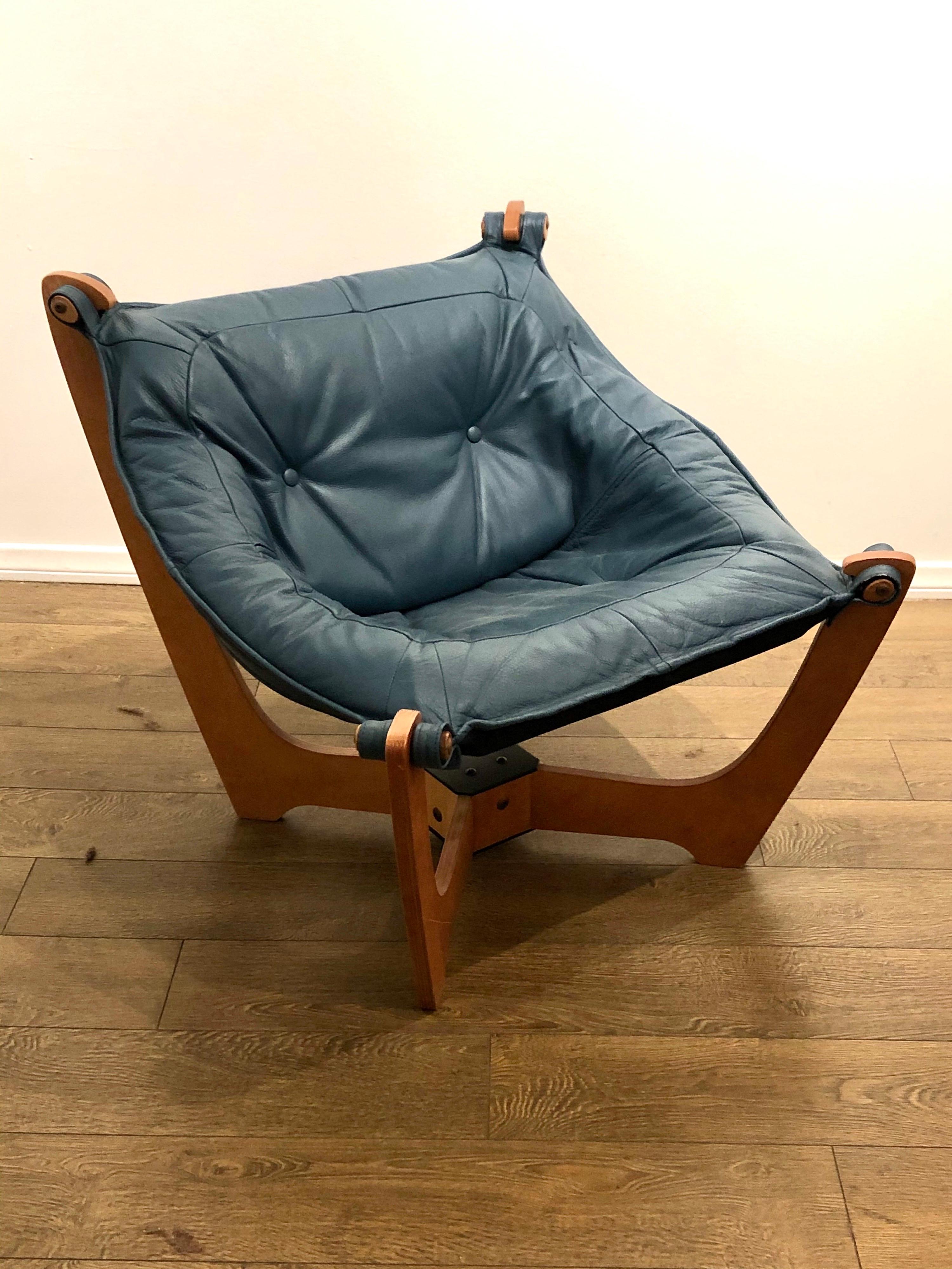 Klassik Luna sling chair is made of birch plywood with a warm natural tone. The four legs come out of a square base on an angle with soft curves leading upward. The seating is covered in turquoise leather and attaches to the legs with knob-like