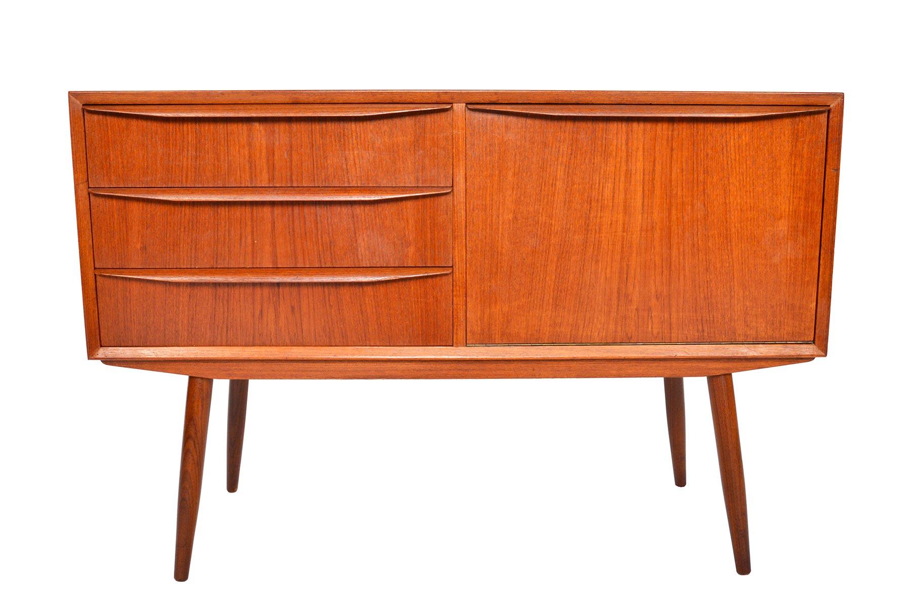 Small and beautifully detailed, this Danish modern teak credenza offers compact storage for the modern home. A bank of three drawers with full profile pulls sits to the left of a drop down cubby. Case stands on canted spindle legs. In excellent