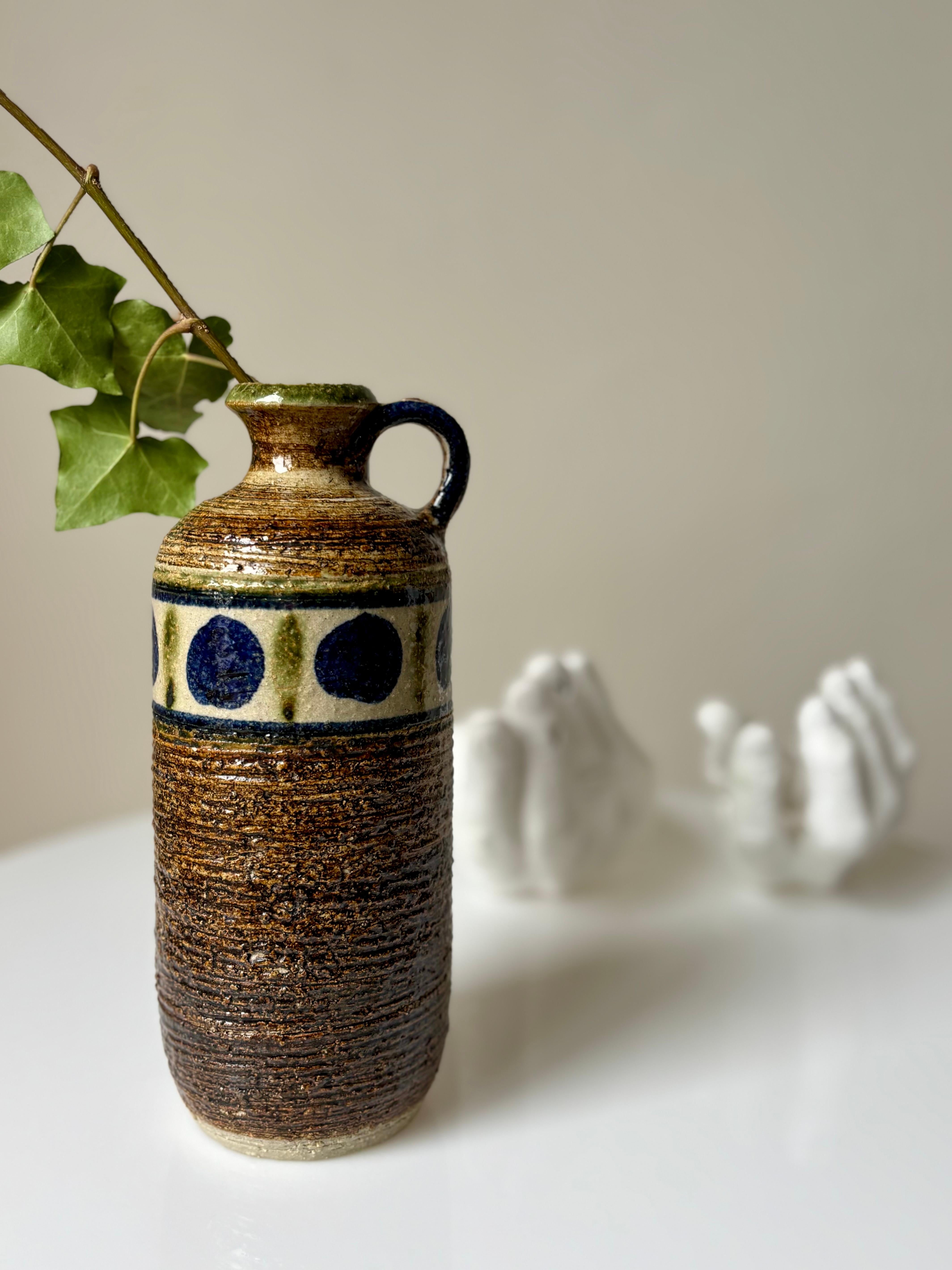 Vaso rustico in gres con smalto marrone, ocra e verde oliva e decorazioni a cerchio blu scuro intorno al bordo superiore. Piccola maniglia sulla parte superiore. Realizzata da Kingo Keramik negli anni '60. 
Firmato sotto la base. 
Bellissime