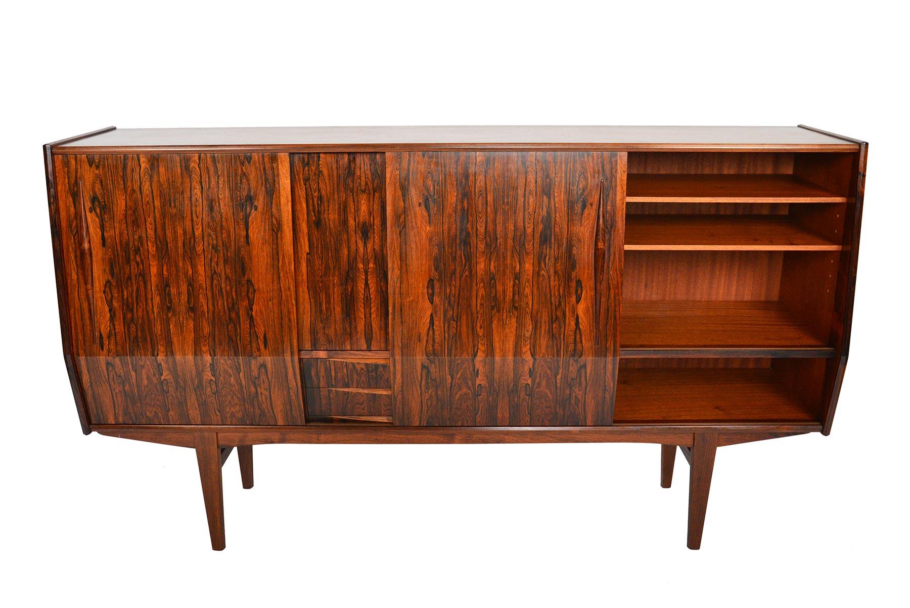 This Danish modern midcentury credenza in rosewood offers exceptional storage in a tall, angular design. Dramatic Brazilian rosewood cathedral grain lines the doors. Four sliding doors open to reveal three bays. Left and right bays offer adjustable