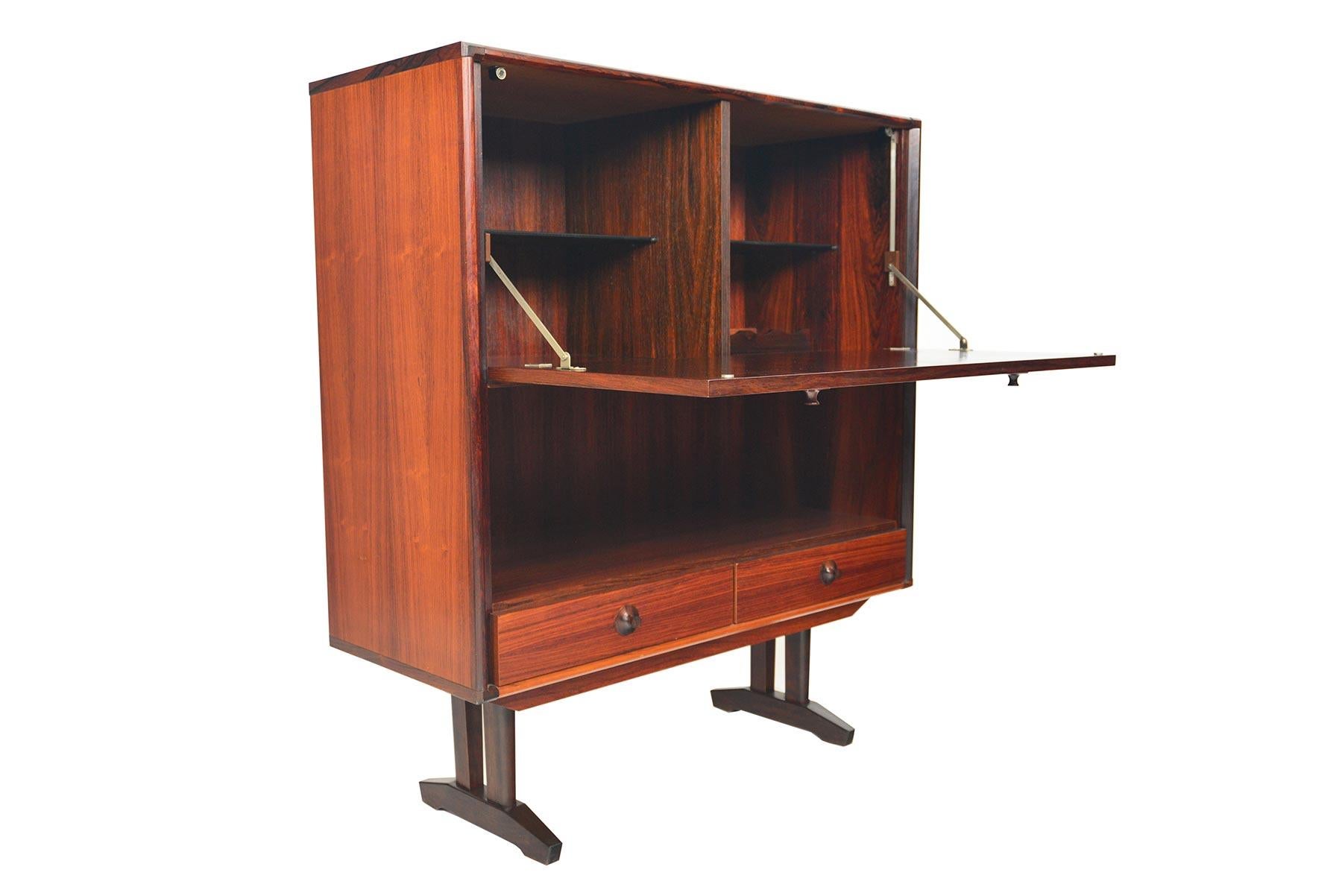 Beautifully cased in Brazilian rosewood, this tall bar was manufactured in Denmark in the late 1960s. A large door with two carved pulls lowers to reveal two bays with black glass shelves and a removable bottle rack. A lower cubby and two lower