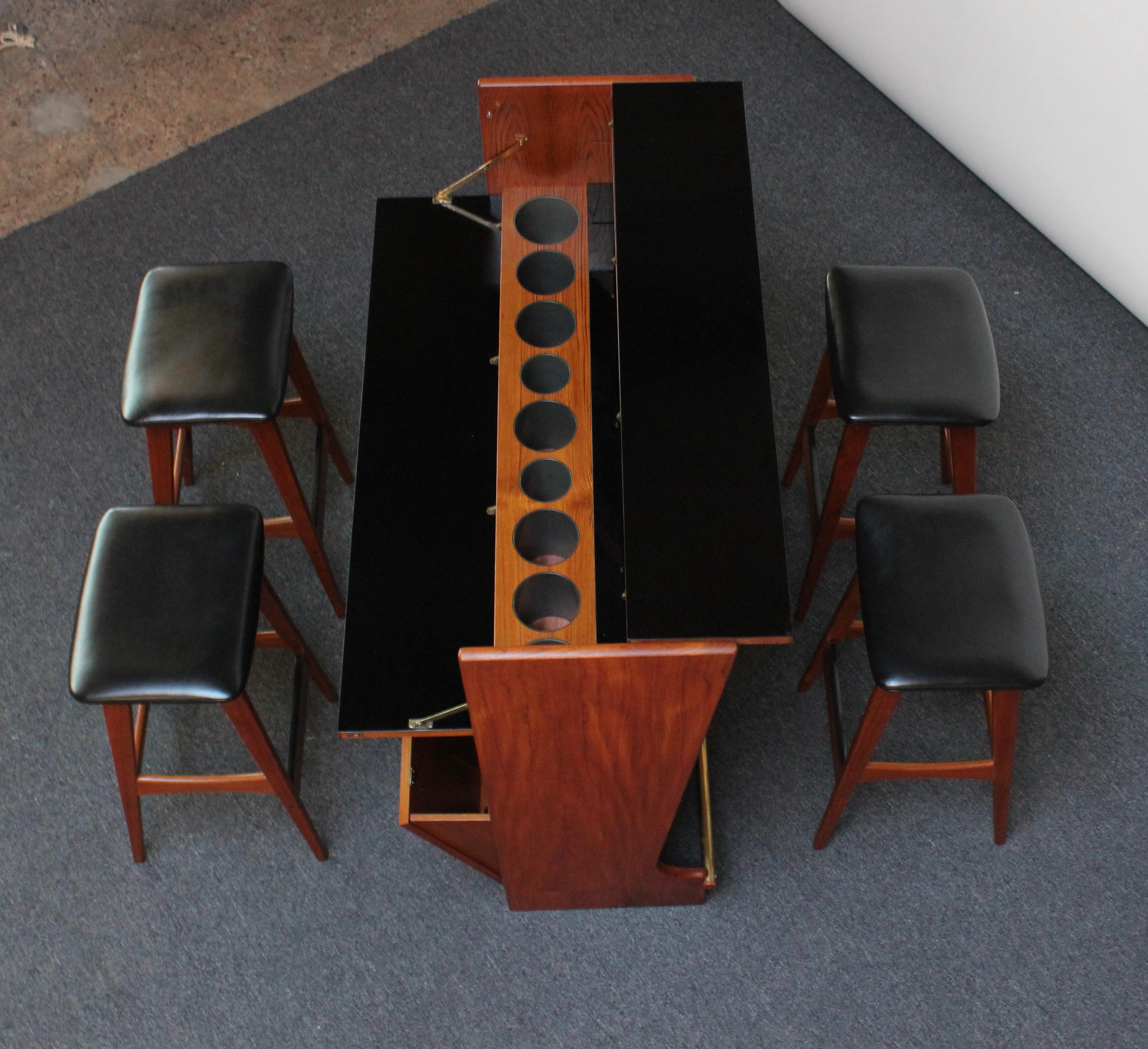 Stylish, functional Danish teak dry bar cabinet designed in the 1960s by Johannes Andersen with four corresponding stools in black leather and teak. 
One side of the bar features an inlaid circular motif and tubular brass footrest, while the other