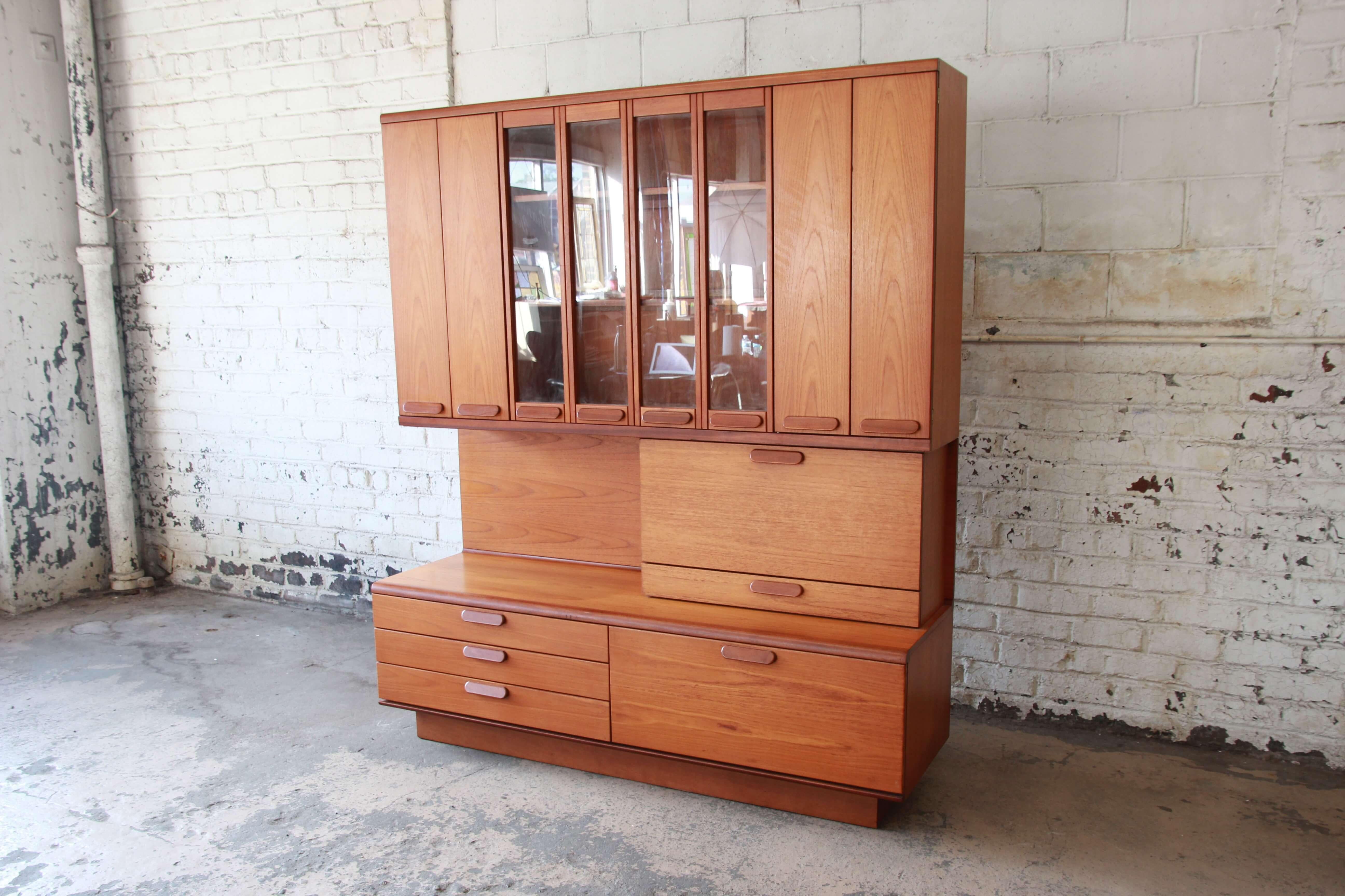 Offering a very nice Danish teak bar wall unit. The cabinet has a great teak wood grain with distinct Scandinavian design. The lower portion has three drawers for storage and a drop-front cabinet space. Just above is a mirrored drop-front cabinet