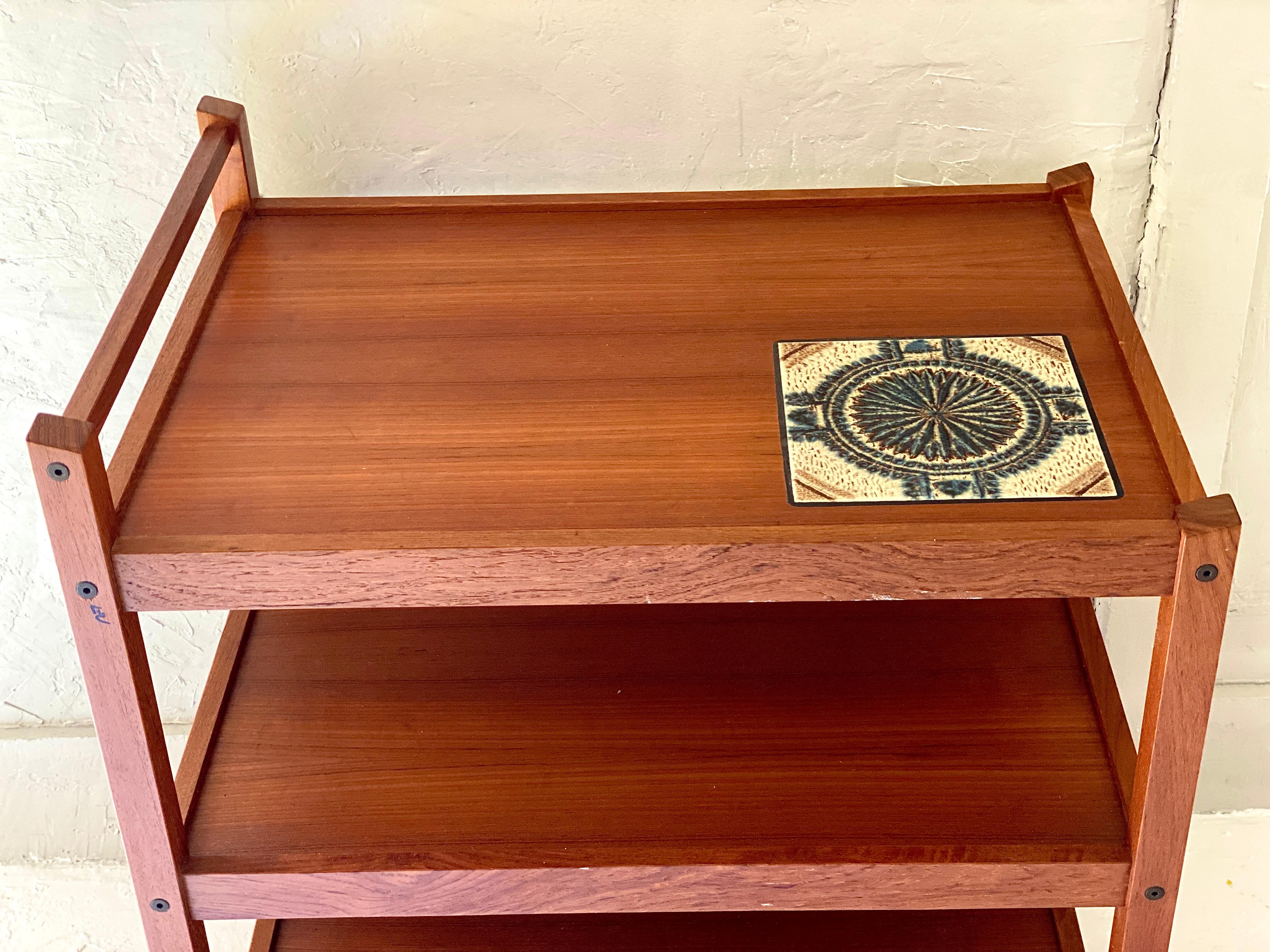 Mid-Century Modern rolling teak bar / tea cart on Casters with Tile on top shelf. This modern style bar cart has 3 shelves and a top handle to easily move from room to room while entertaining guests. The top tile can be used for putting a bottle of