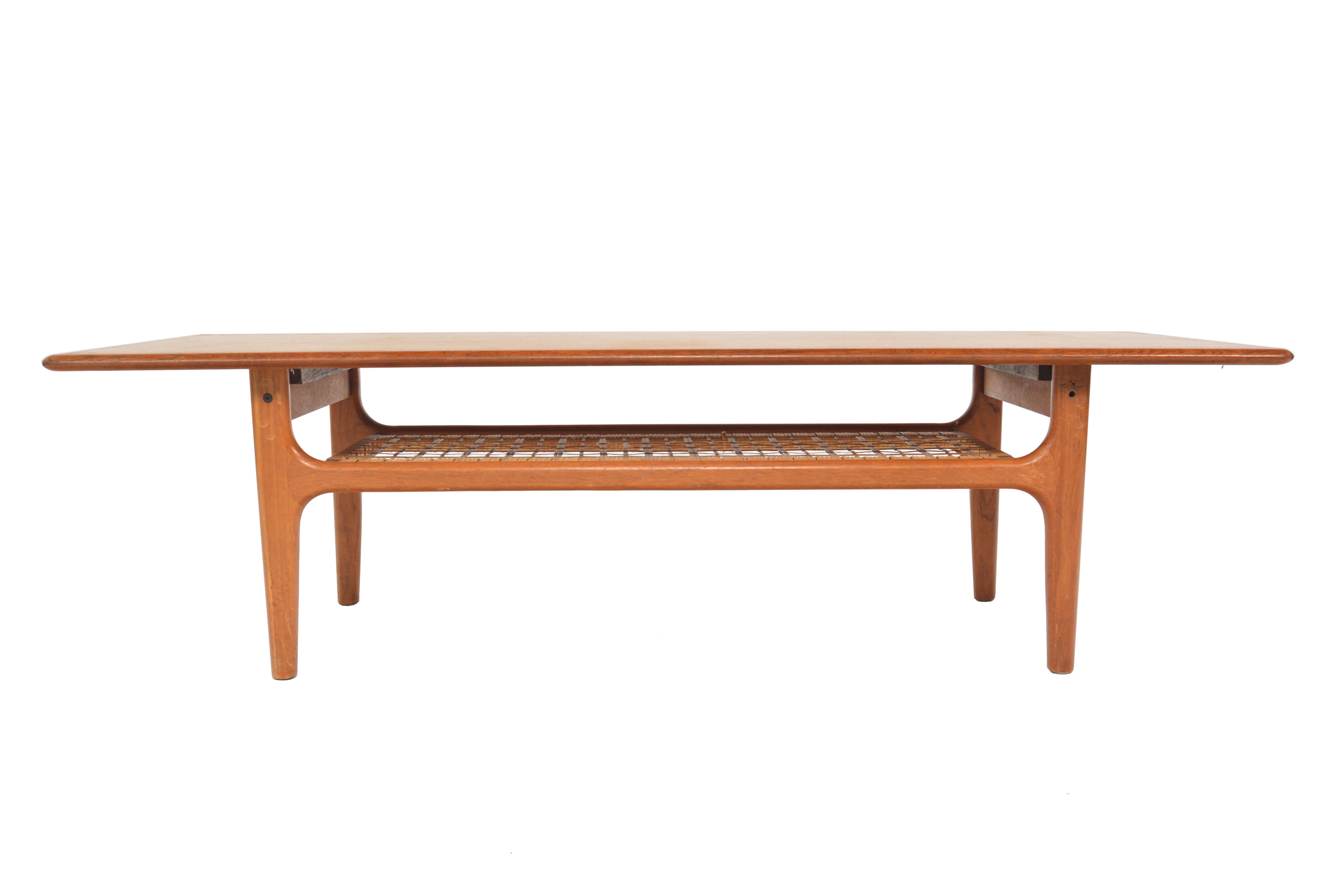 This Danish modern midcentury surfboard coffee table in teak by Trioh is beautifully designed. The rectangular teak slab features a softly sculpted banding. An original cane rack underneath the table offers storage. In excellent original