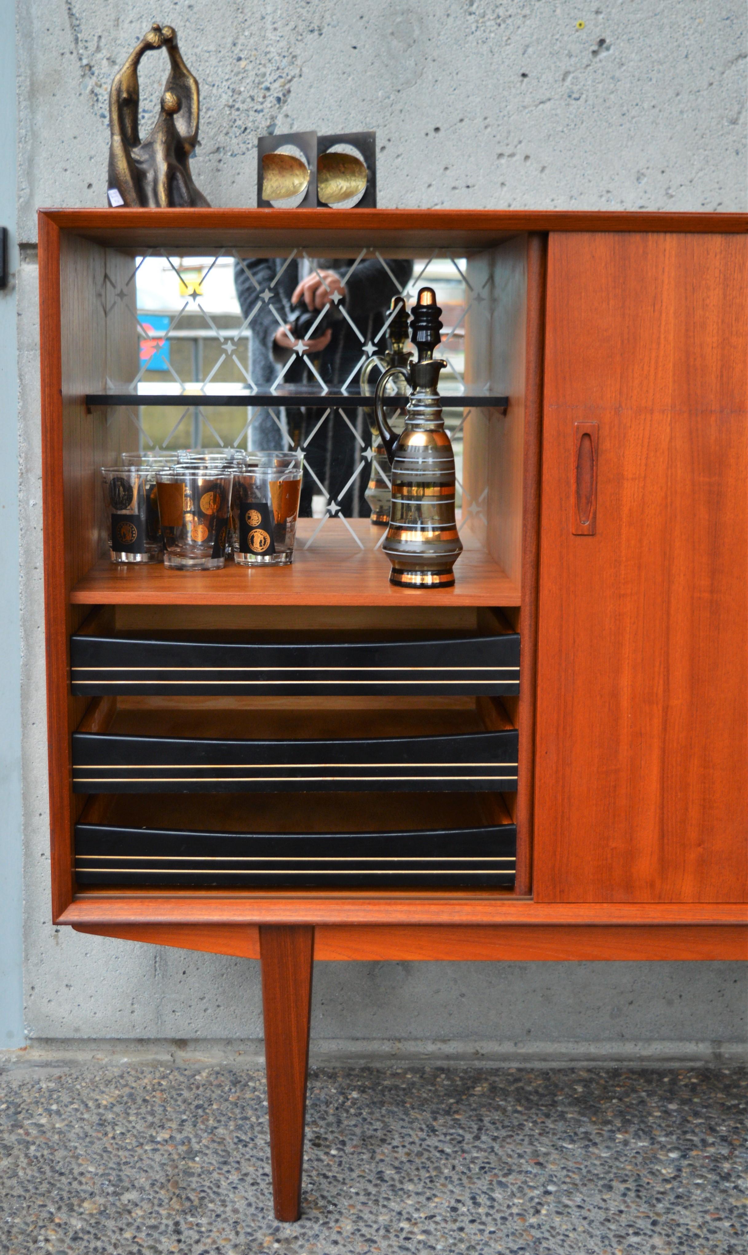 This hot, Danish Modern teak taller credenza/sideboard offers a fantastic amount of storage on a relatively small footprint. Raised up on tall sword blade legs, it features a beveled from molding detail, and 3 sliding doors with ovoid inset door