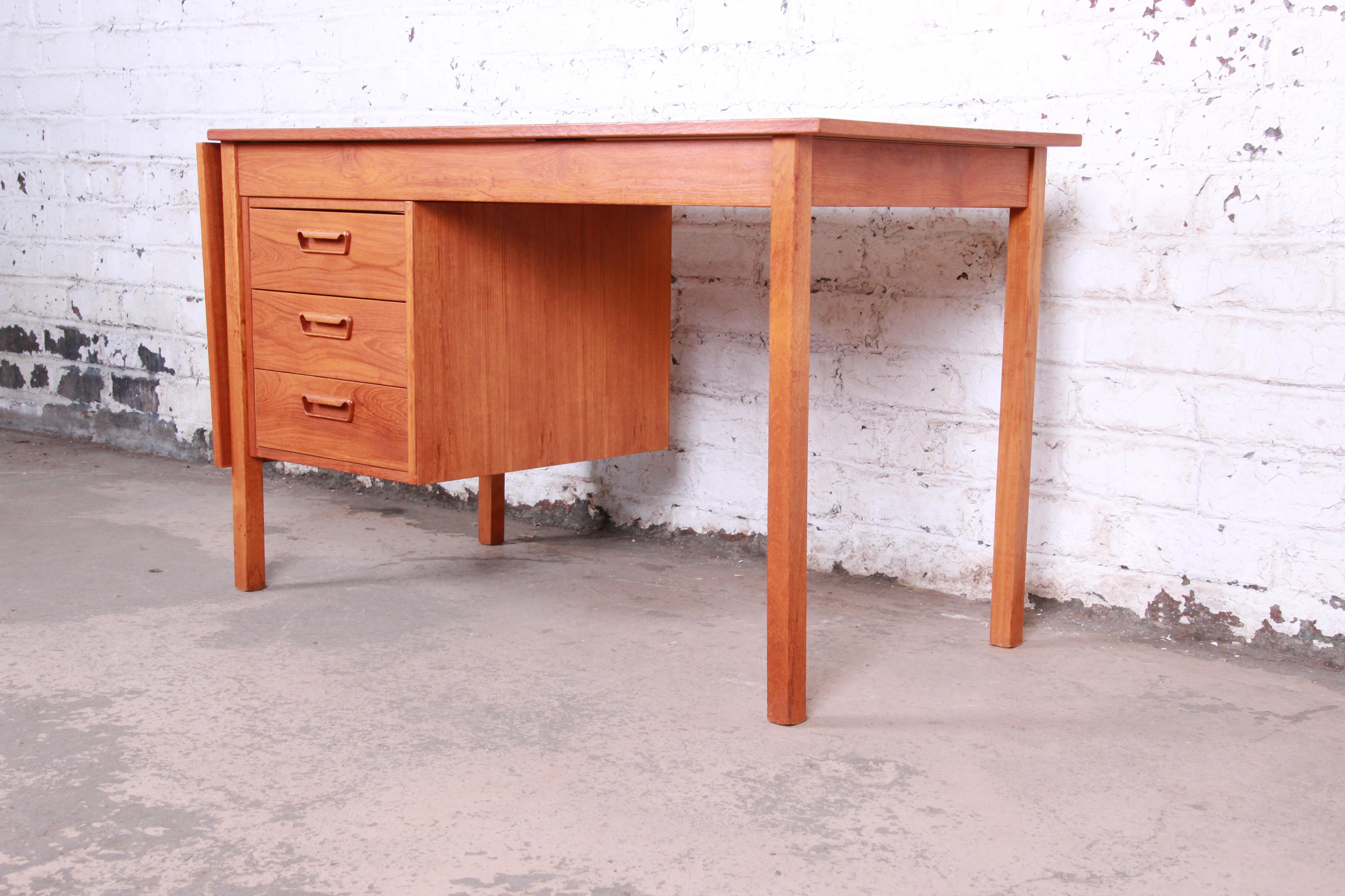 A gorgeous Danish modern teak drop-leaf desk. The desk features stunning teak wood grain and sleek, minimalist midcentury Danish design. It offers good storage with three drawers, and the leaf flips up to extend the table top. The desk is finished