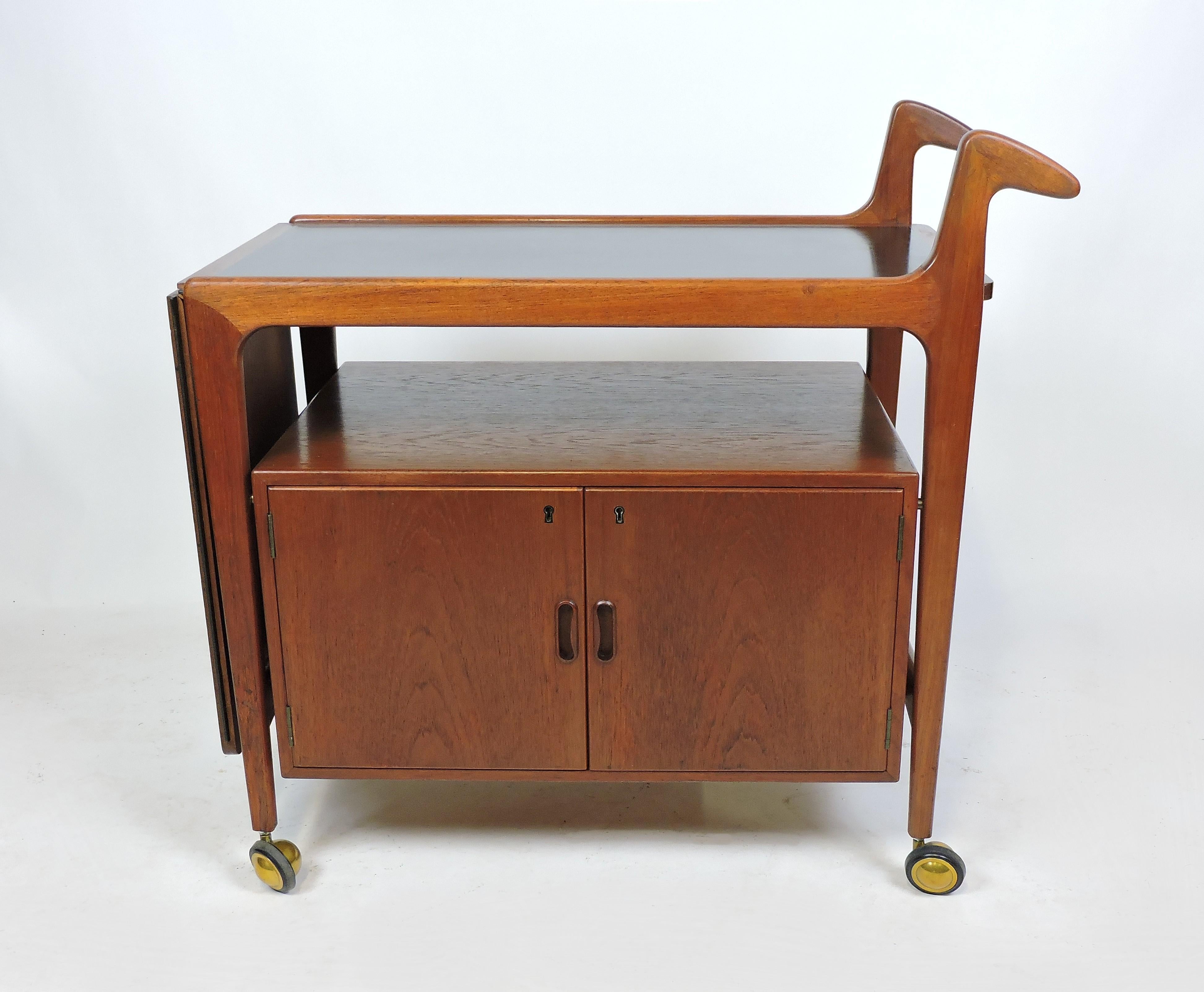 Handsome and practical teak bar cart made in Denmark by Falster Mobelfabrik. This cart can be wheeled around for serving using beautifully sculpted handles. It also has a locking cabinet with an interior adjustable shelf and a black formica surface