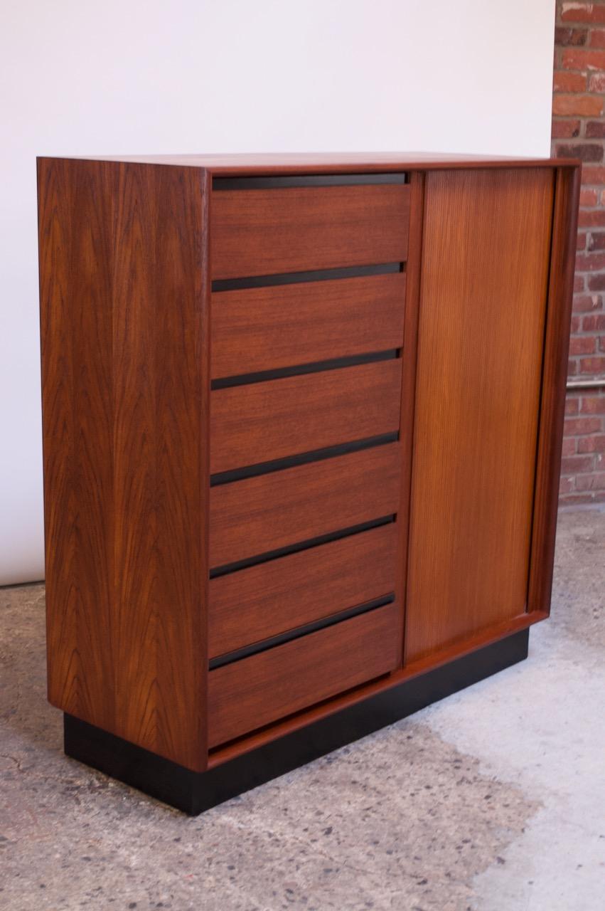 Danish modern gentleman's chest / highboy in teak supported by an ebonized plinth base designed by Art Furn and manufactured by Danflex Systems, (Denmark, 1960s). Six drawers on the left side, seven on the right concealed by a tambour door.