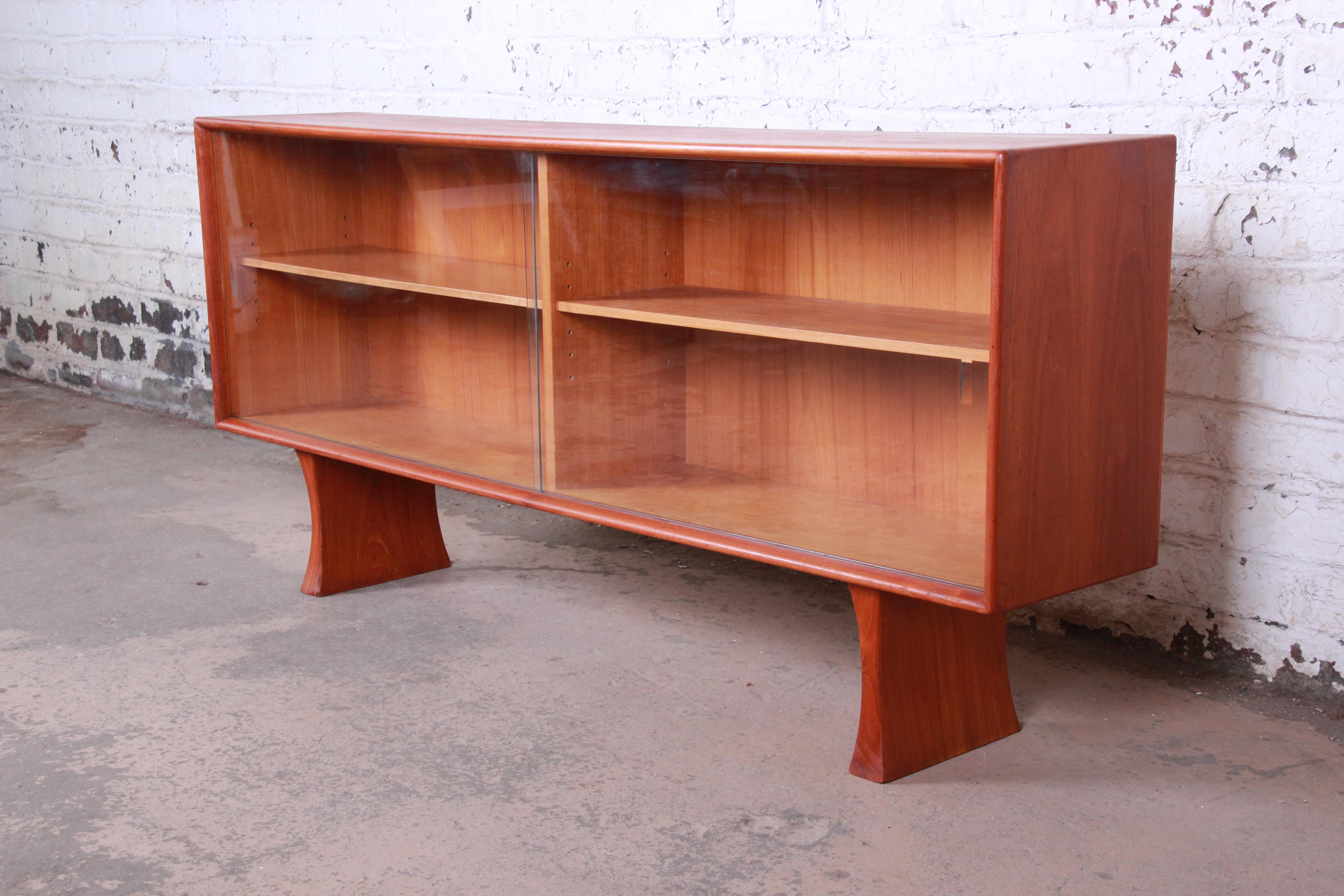 A stunning midcentury Danish modern teak glass front bookcase or credenza. The credenza features beautiful teak wood grain and sleek Danish design. It has a floating design, with two sculpted solid teak legs and a teak case that is finished on all