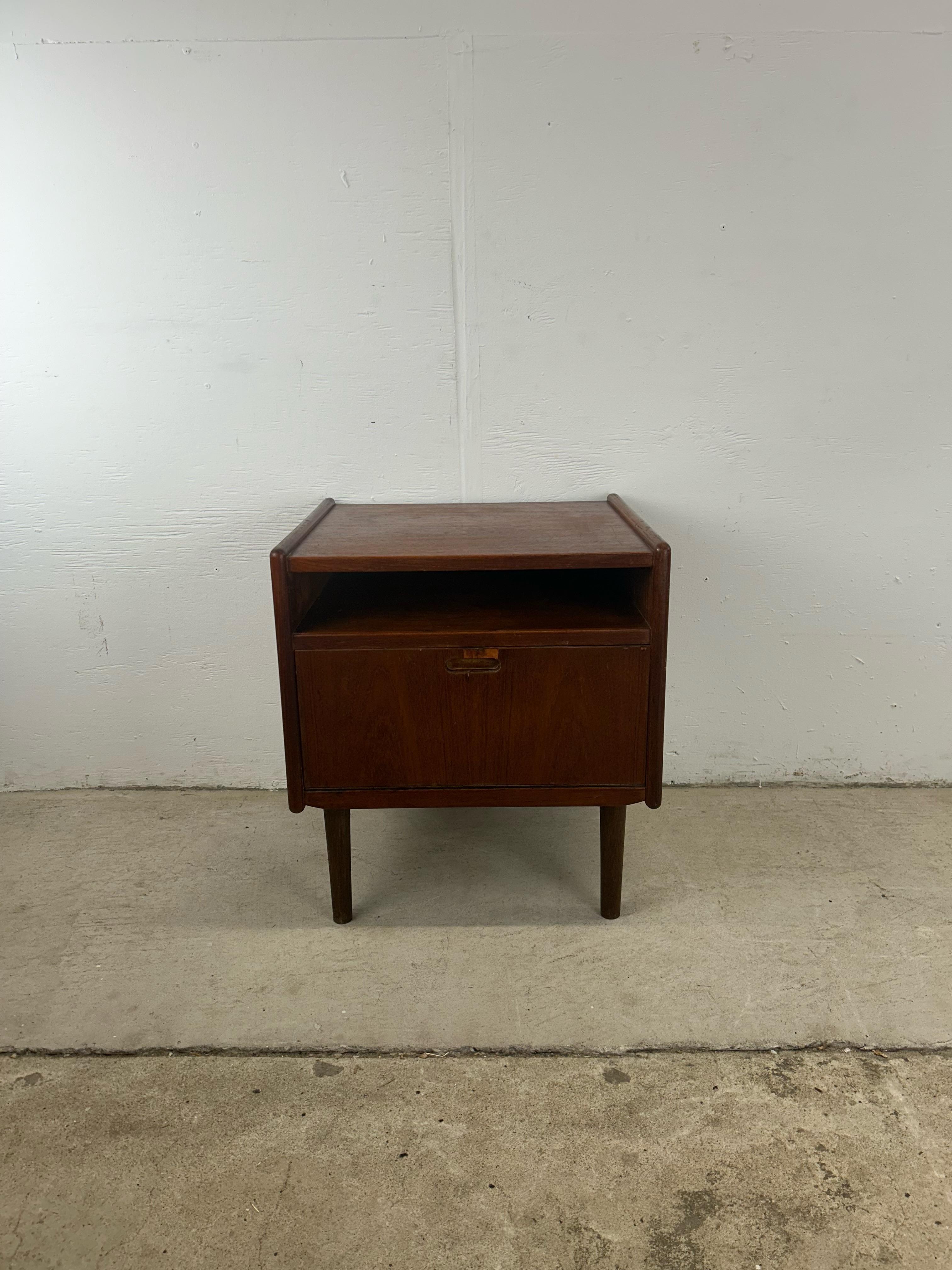 This danish modern nightstand features solid teak construction, removable tapered legs, and drop front design with interior storage.
 Dimensions: 19.5w 17.75d 21.5h

Condition: Original cabinet pull was damaged and lost with time. There is a large