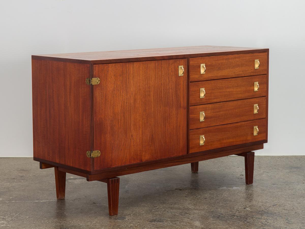 Petite teak credenza, designed by Peter Lovig Nielsen for Dansk Designs. Eye-catching brass hinges and pulls add an elegant touch to this Danish Modern cabinet. Hand-carved legs are constructed with interlocking finger joints that elevate the whole