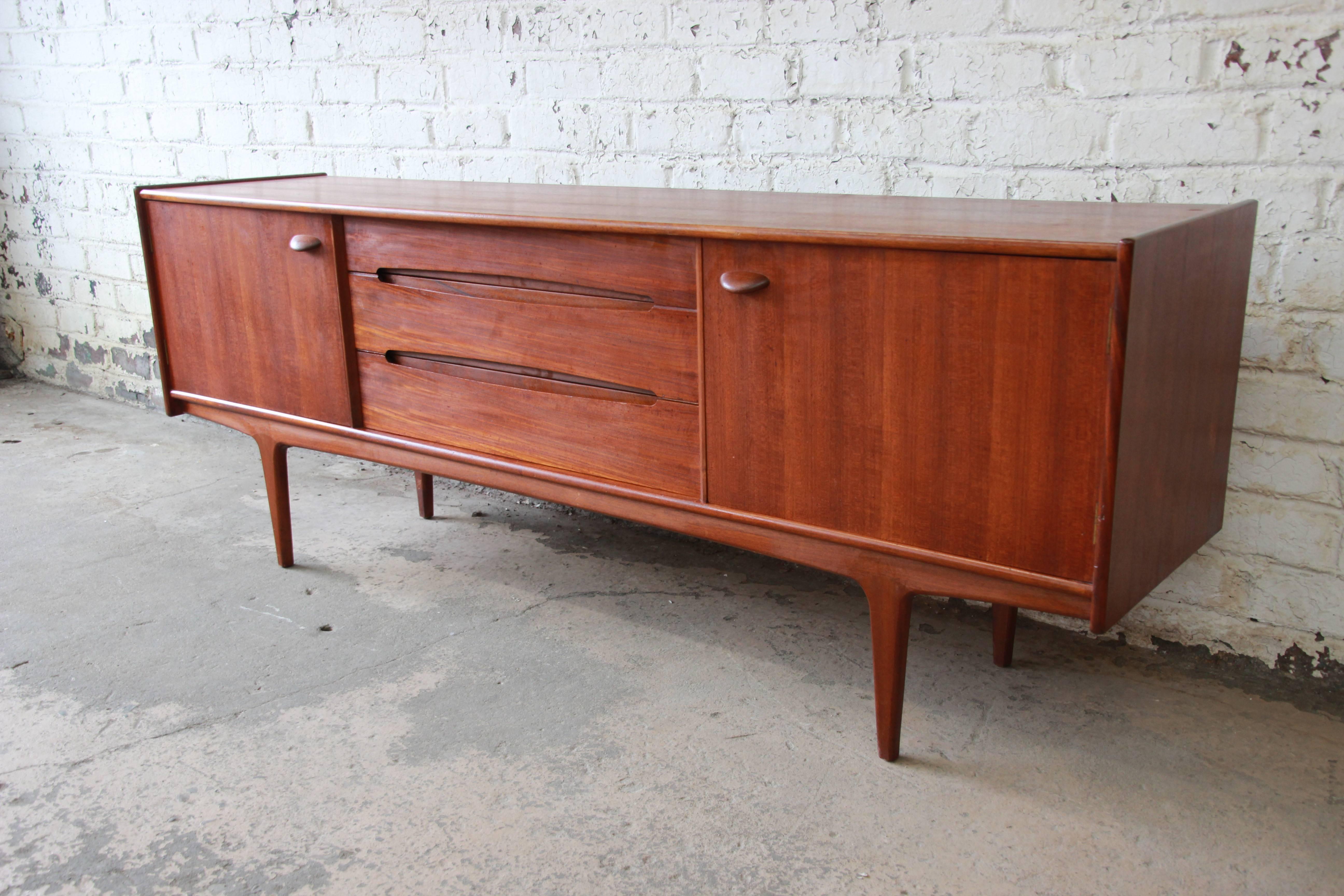 A stunning Danish modern style teak sideboard credenza by Younger. The credenza features gorgeous teak wood grain and sleek midcentury design, with a uniquely designed slightly curved front edge. It offers ample room for storage with three