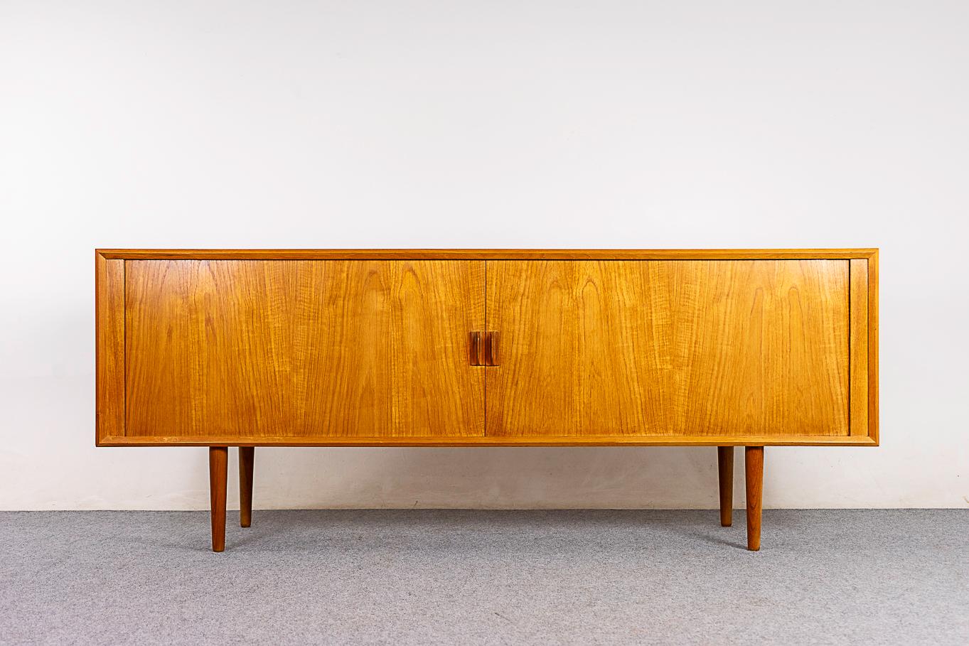 Teak mid-century sideboard, circa 1960's. Beautiful bookmatched veneer, solid wood edging and sleek tapered legs. Magical disappearing tambour doors slide open to provide seamless access to a spacious interior. Height adjustable shelving and slender