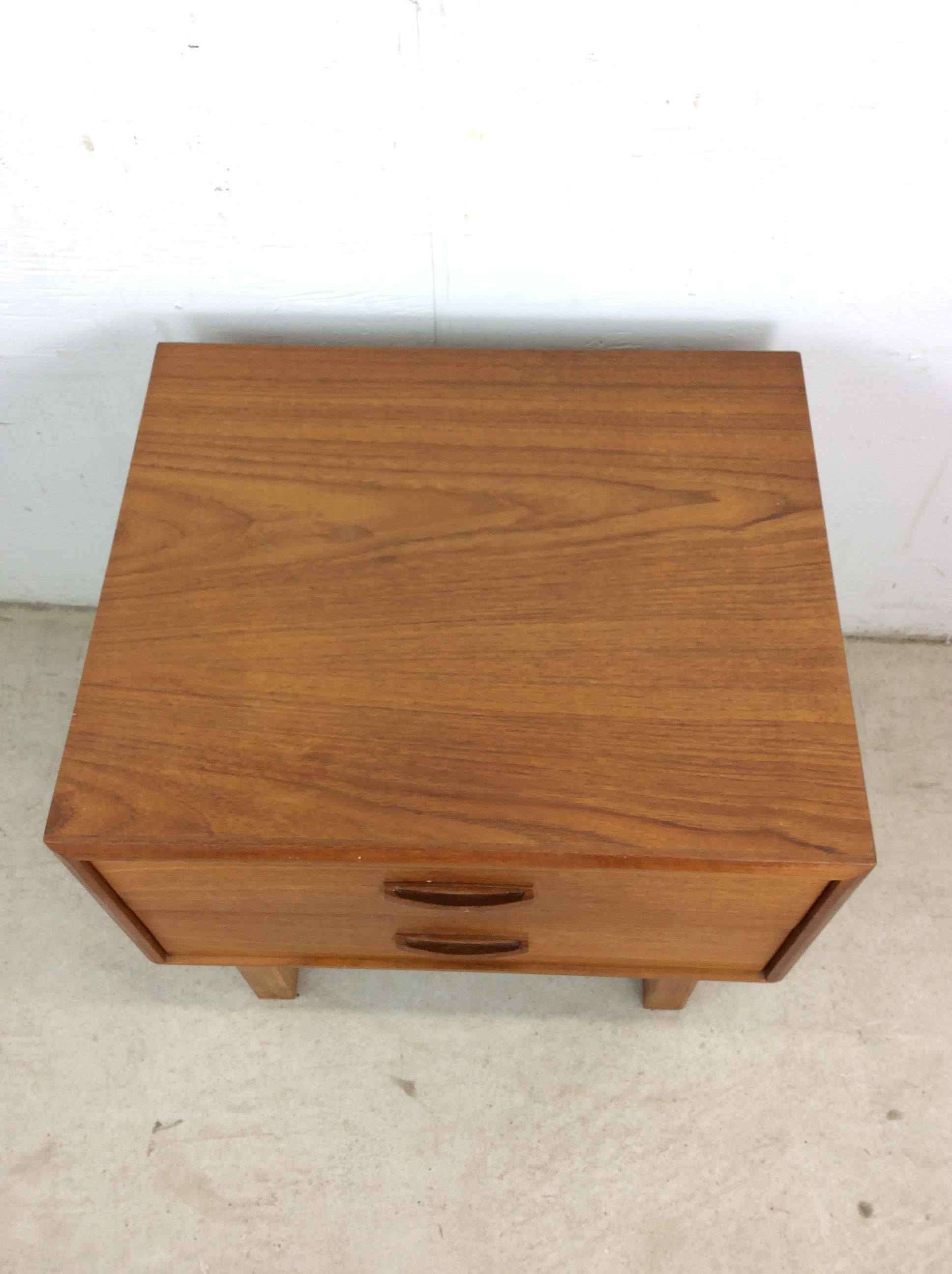 This Danish modern teak nightstand features pressed wood construction, teak veneer with original finish, two drawer design with carved wood pulls, and tall tapered legs.   Matching chest of drawers with mounted mirror and highboy dresser available