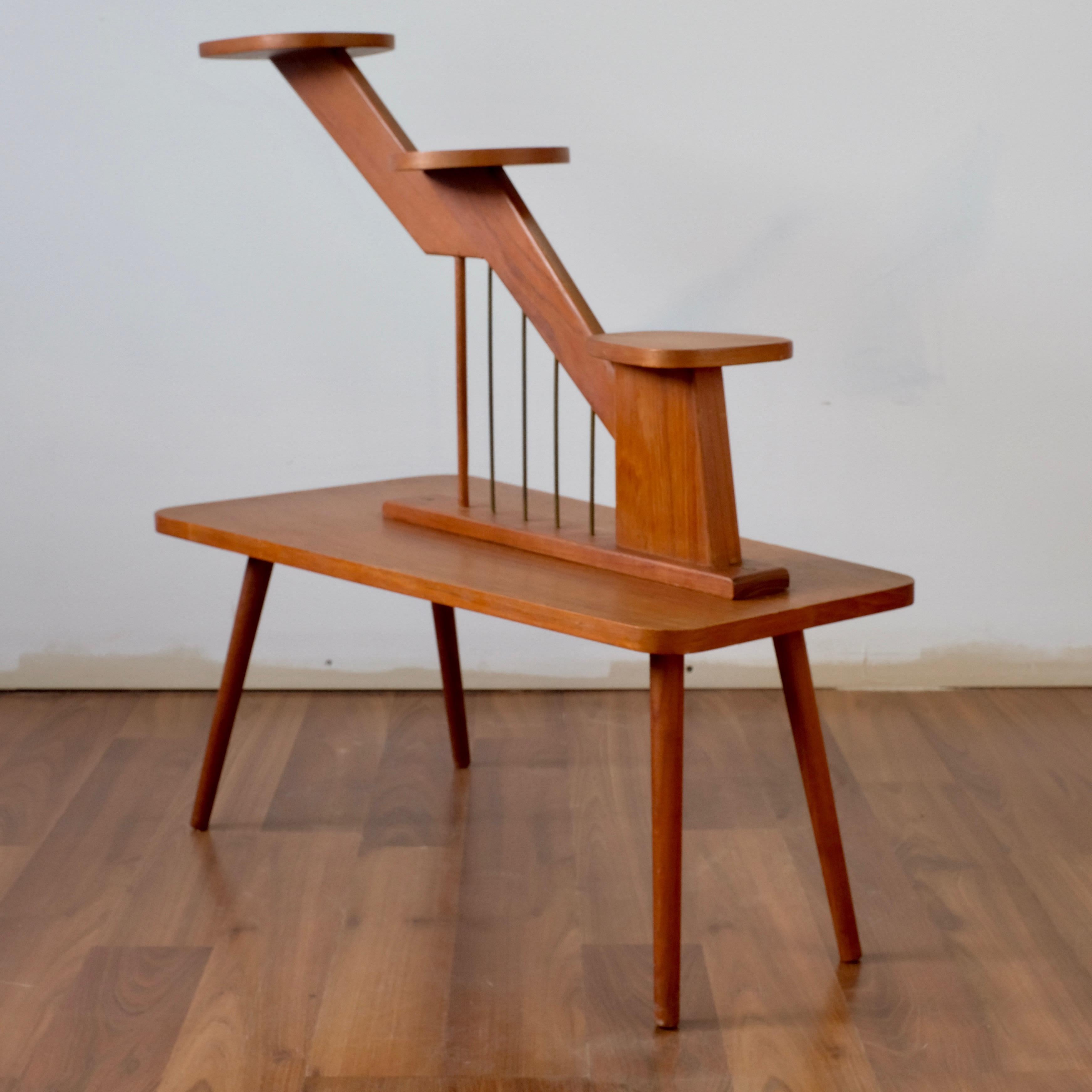 Three-tiered plant stand in solid teak with distinctive harp-like vertical supports in patinated metal. 

The four tapered legs screw in to angled brass brackets. Attached to the bottom of the base.