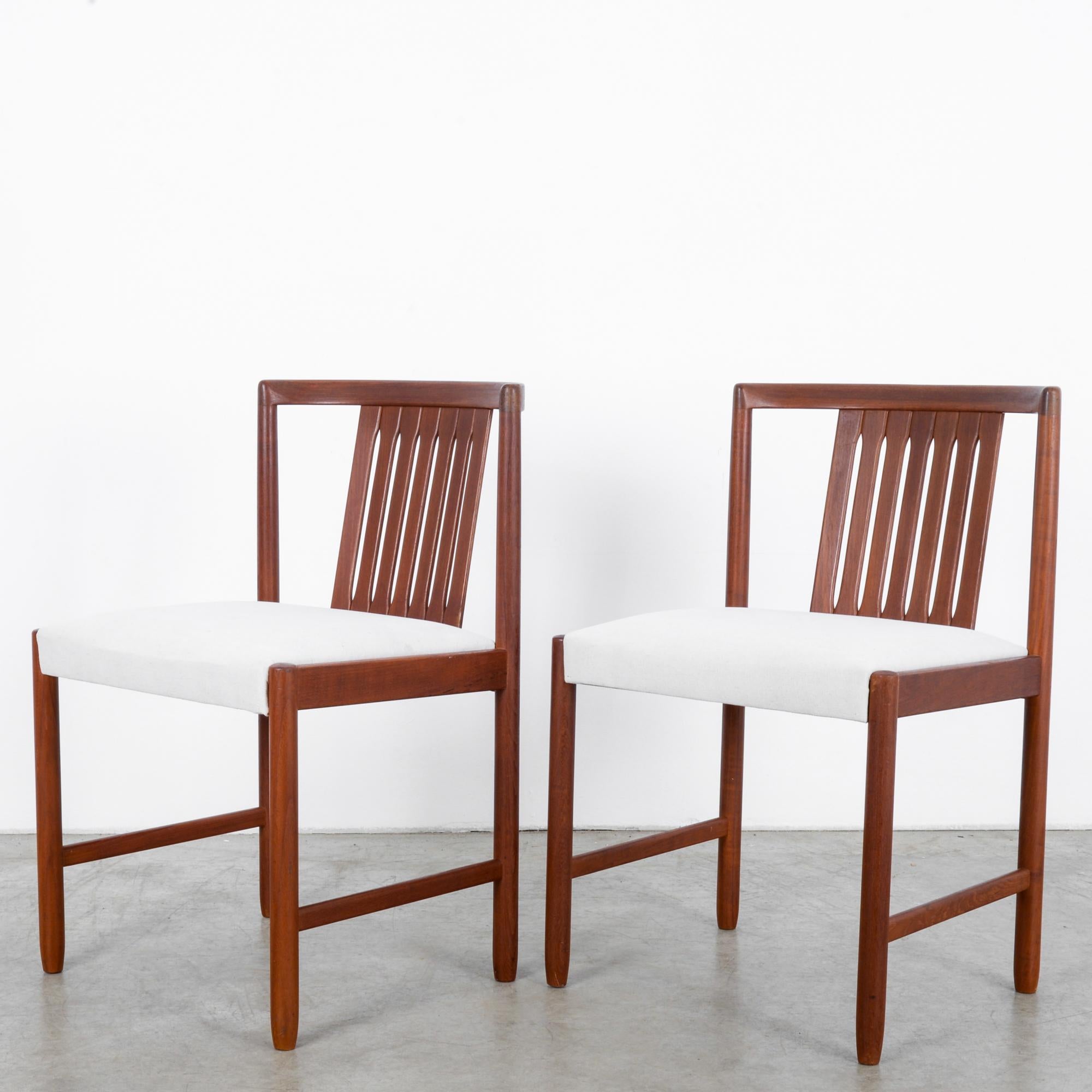 This pair of teak chairs was made in Denmark, circa 1960. From the front, the chairs appear to have an angular structure, and the side profile reveals the curved top rails and angled slats of the back. The ivory white upholstered seats highlight the