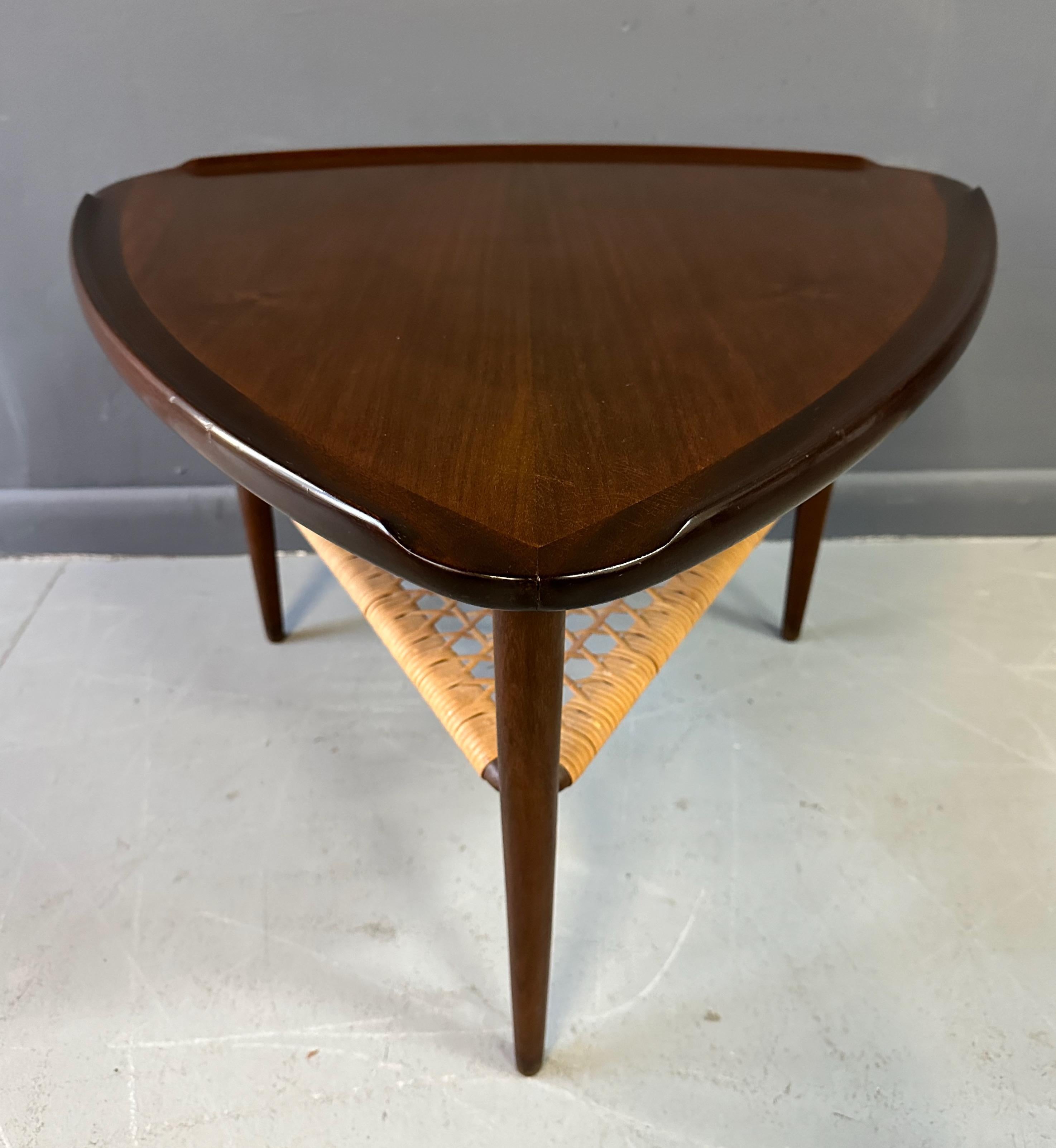Mid Century triangular occasional guitar pick side table in walnut designed by Poul Jensen imported by Selig, Denmark. This piece features a unique raised edge top and lower tier magazine shelf made of woven cane.