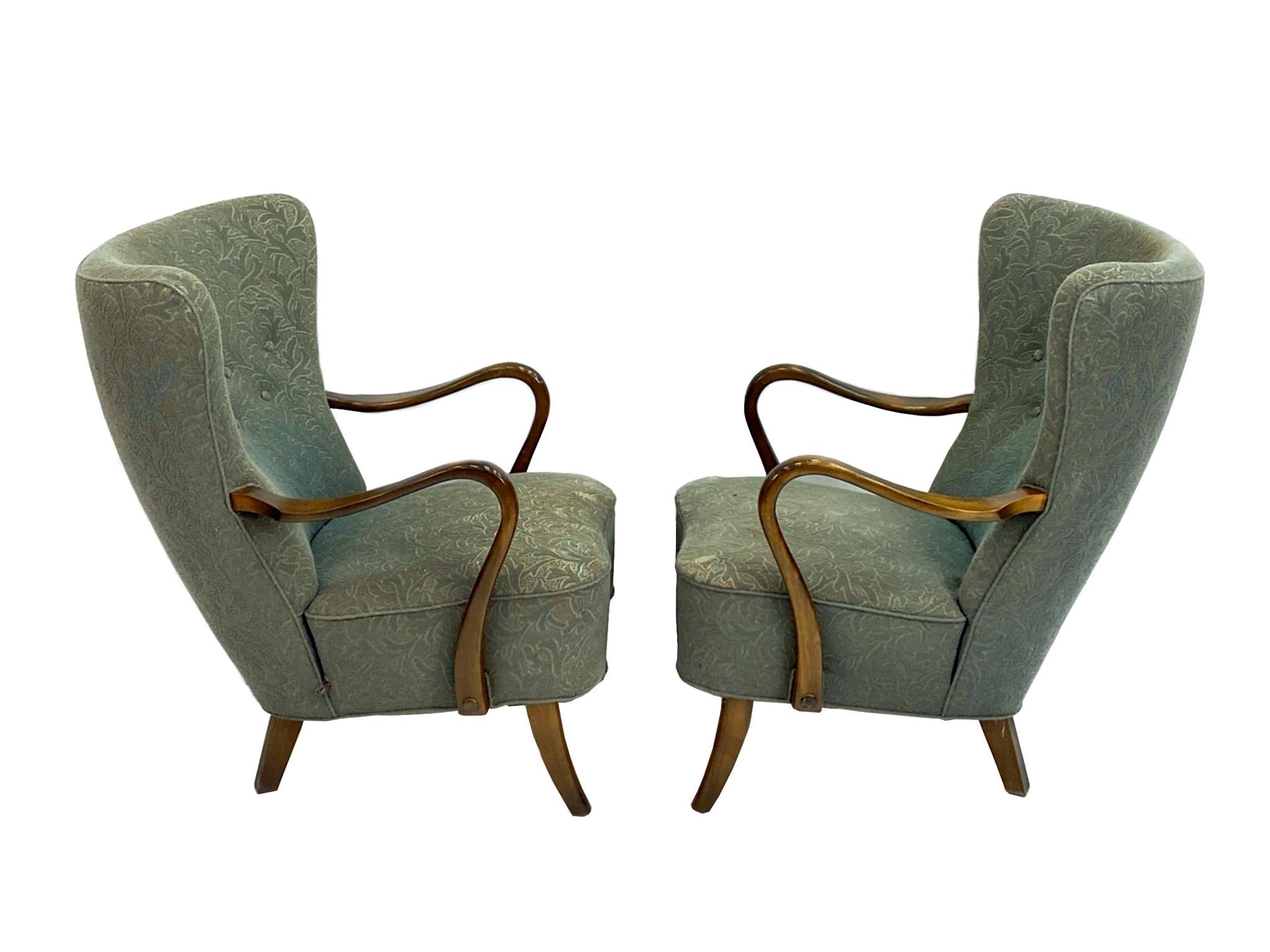 Fabriqués dans les années 1950, ces fauteuils à oreilles se composent d'une structure en bois de hêtre et d'un revêtement en tissu à motifs vert mousse. Les formes incurvées caractérisent le design, ce qui se voit dans les bras joliment inclinés et