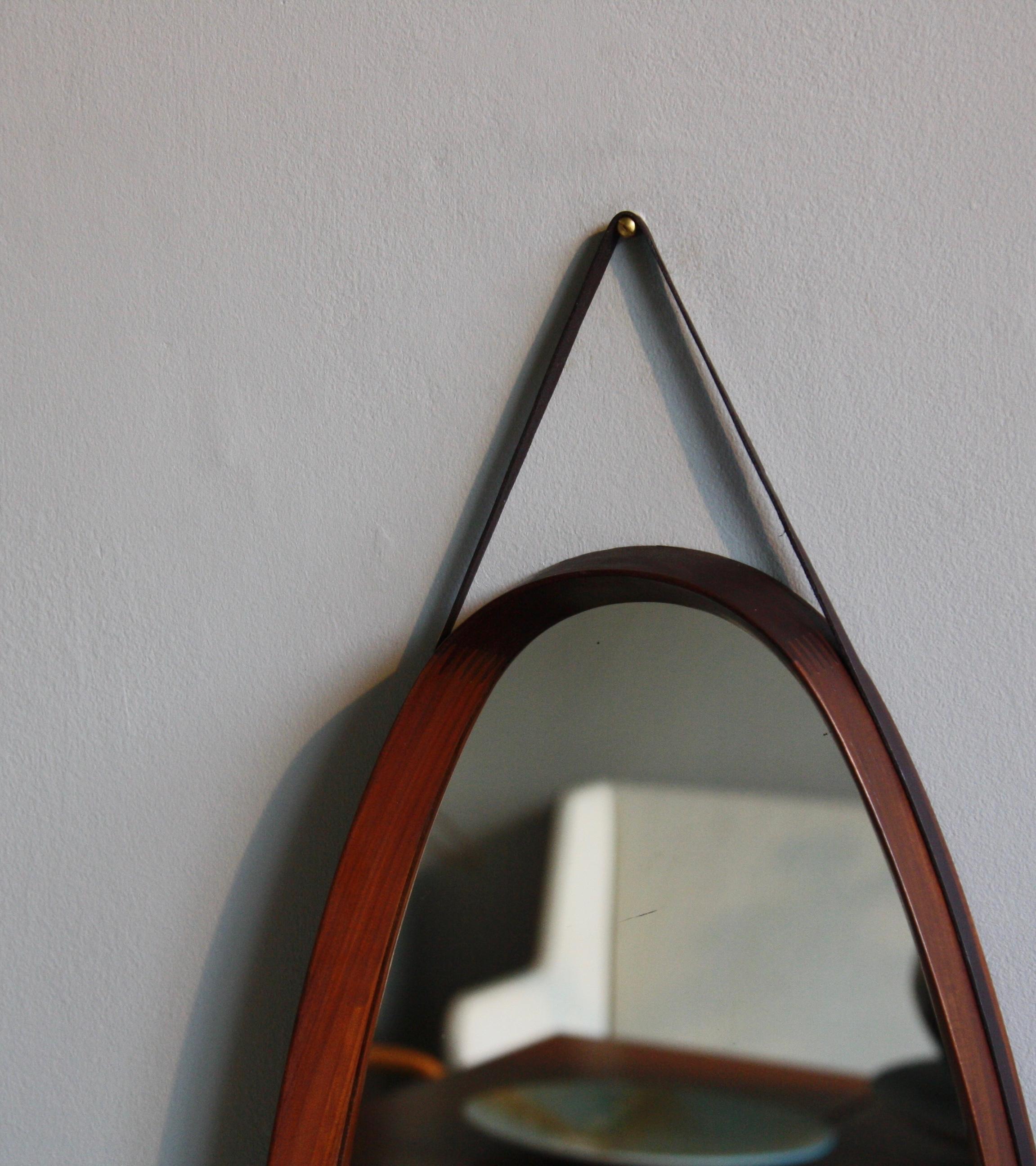 An oval shaped vintage wall mirror with leather hanging strap, made in Denmark, circa 1950. The mirror is made of sections of solid teak wood which are joined together using finger joints. This exposed construction, the tan colored leather strap and