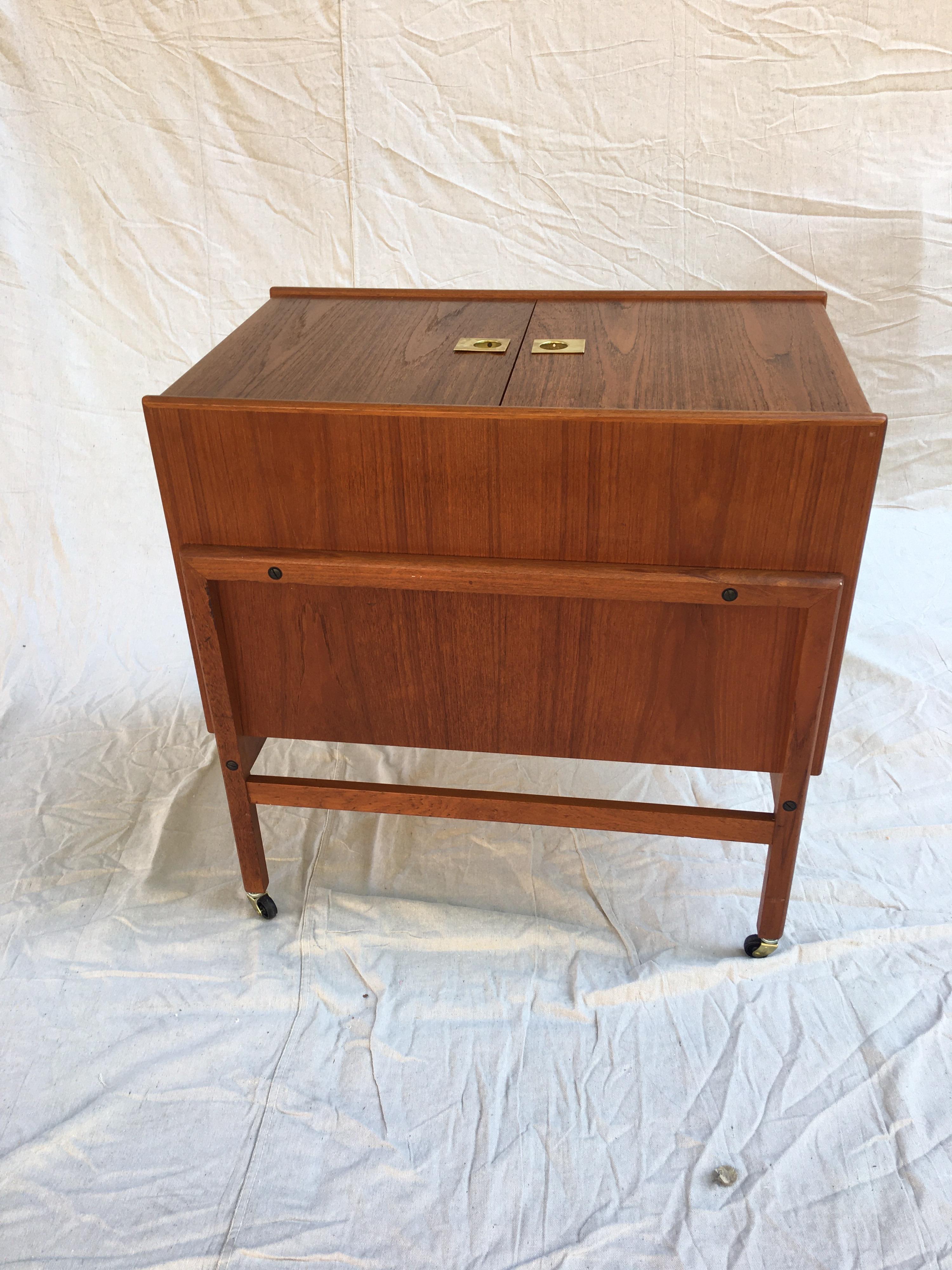 Nice compact rolling bar cabinet, top 2 doors open to reveal bottle storage and lids function as a work surface. Teak with brass accents.