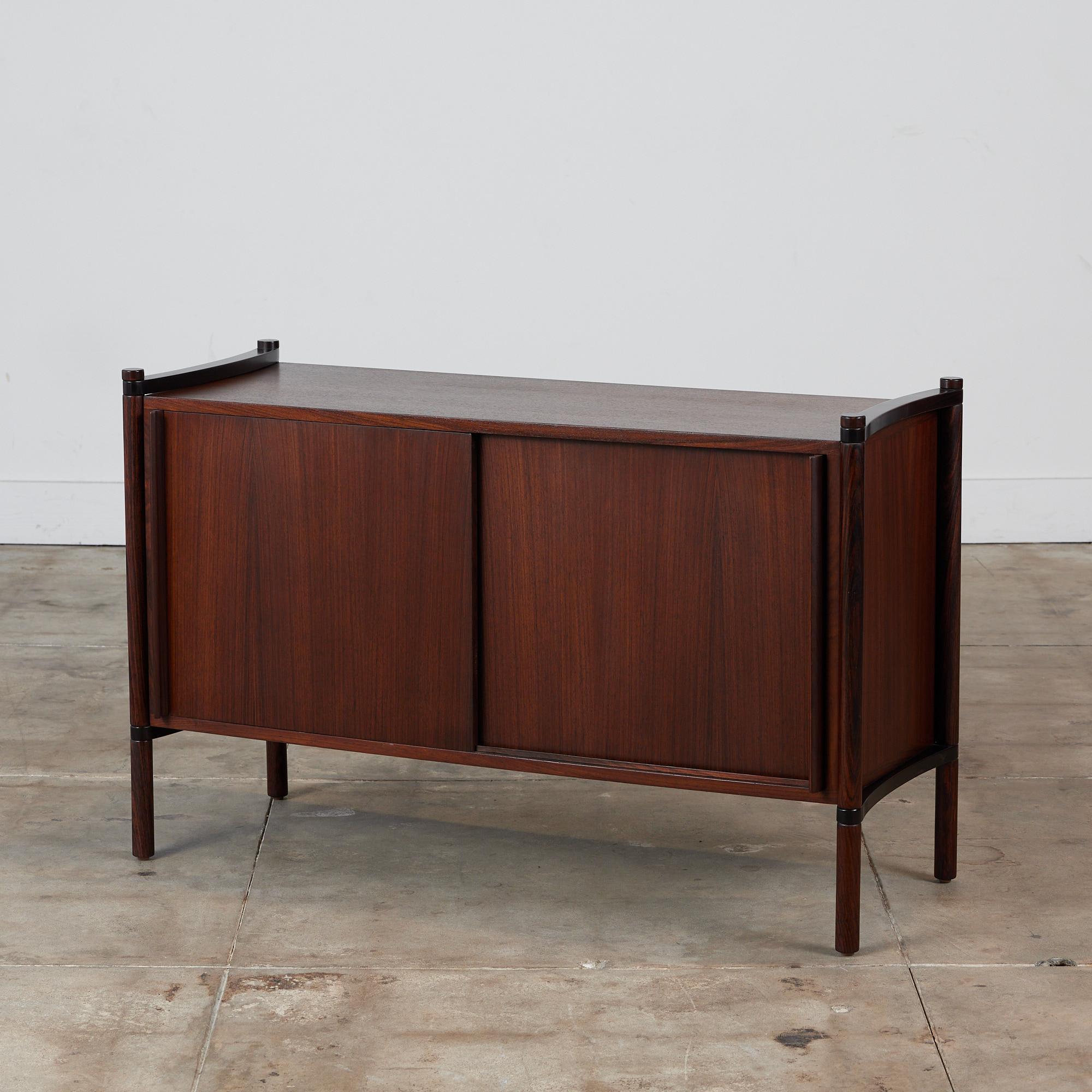 Sliding two door Rosewood credenza featuring two shelves supported by four cylindrical dowels that run the height of this case piece. Each dowel extends slightly above the unit and below to the floor. The credenza has a bowed wood detail on the top