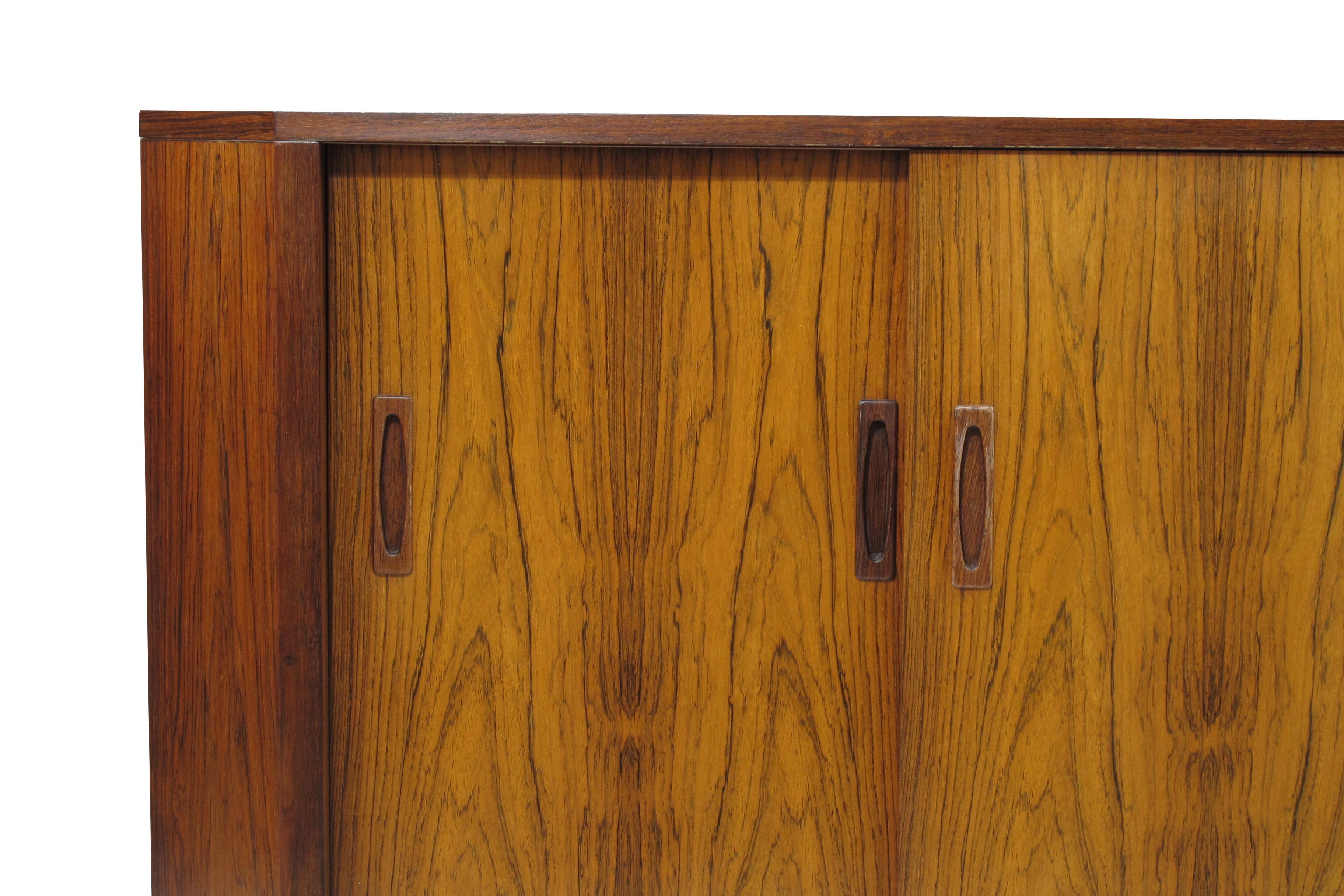 Midcentury low corner cabinet manufactured in Denmark. Cabinet crafted of rosewood with bookmatched grain and two sliding doors featuring carved inset pulls. Interior has two adjustable shelves. Fully restored. Leg clearance 9.63