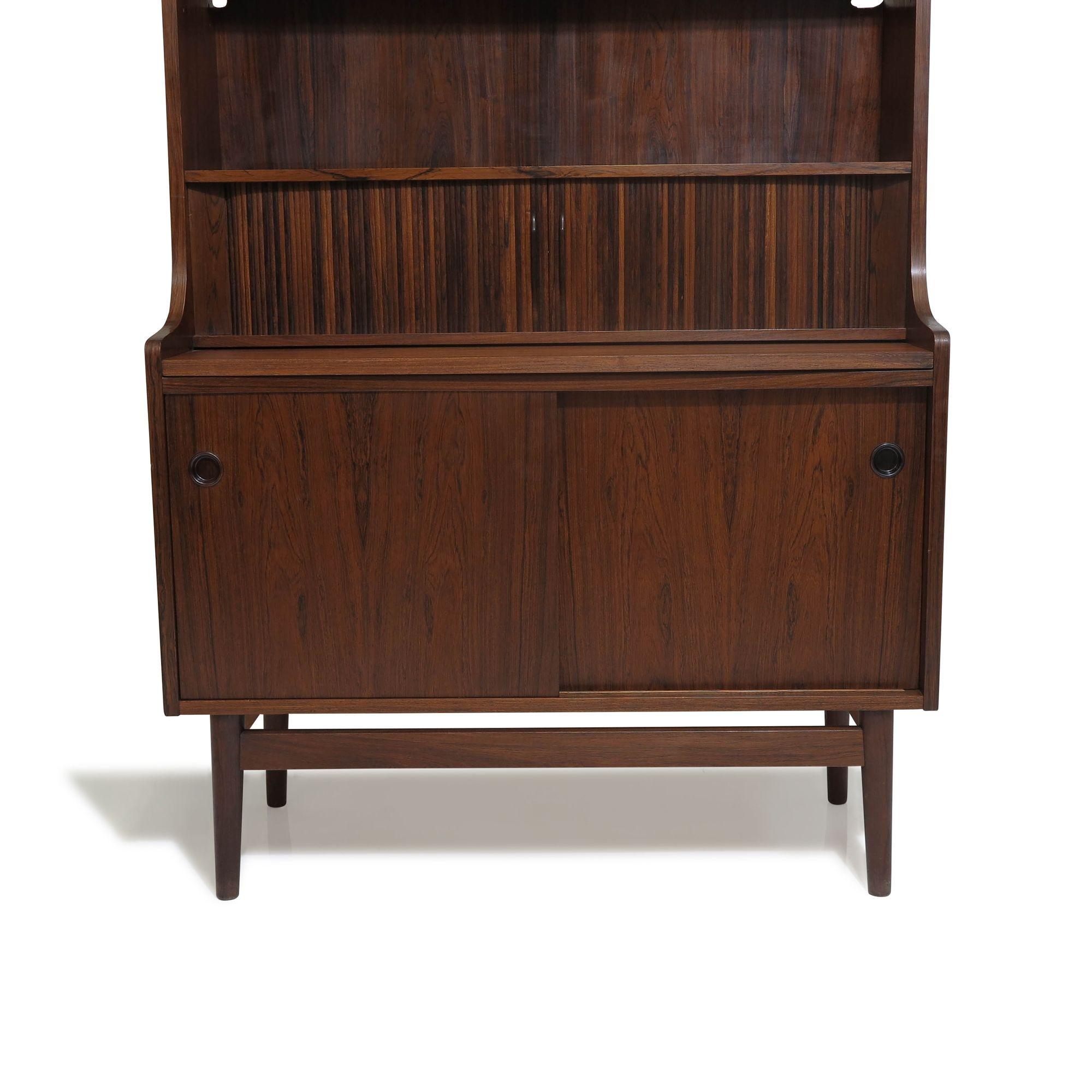 Mid century secretary desk and bookcase crafted of Brazilian rosewood with pull-out writing surface over a small cabinet with sliding doors. Mid-section has pair of solid rosewood tambour doors opening to reveal three small drawers and mail