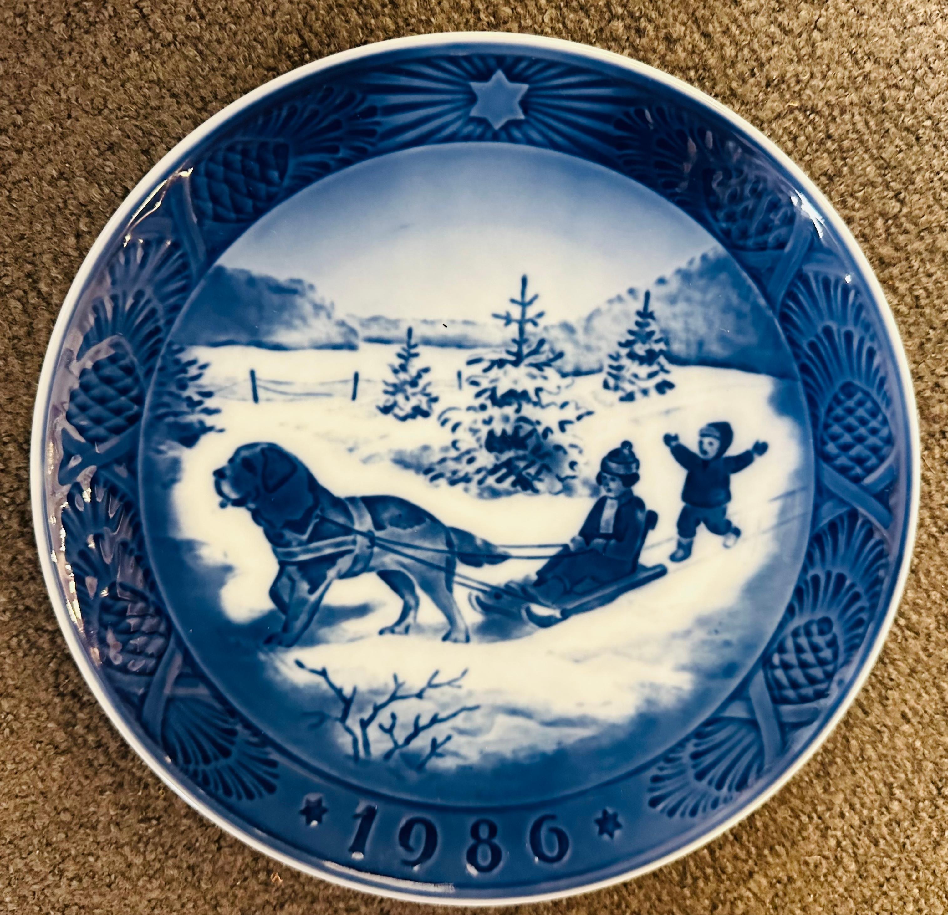 1986 Danish Royal Copenhagen Christmas plate entitled Christmas Holidays (Juleferie). Designed by Sven Vestergaard depicting a scene showing children playing in the snow with the older child sitting on a sledge being pulled by a Saint Bernard dog