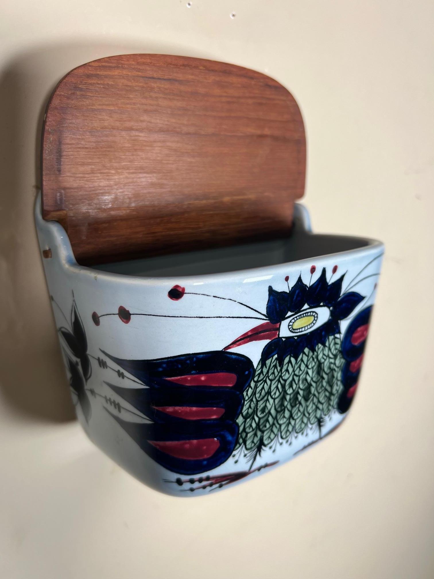 Danish ceramic wall mounted salt box by Royal Copenhagen. Original stamp at the back. With wooden lid. Fajance (tin-glazed earthenware ceramic). Numbered 132/261
Very good condition. Small mark on lid. Looks like a small burn mark. 
Dimensions: 6