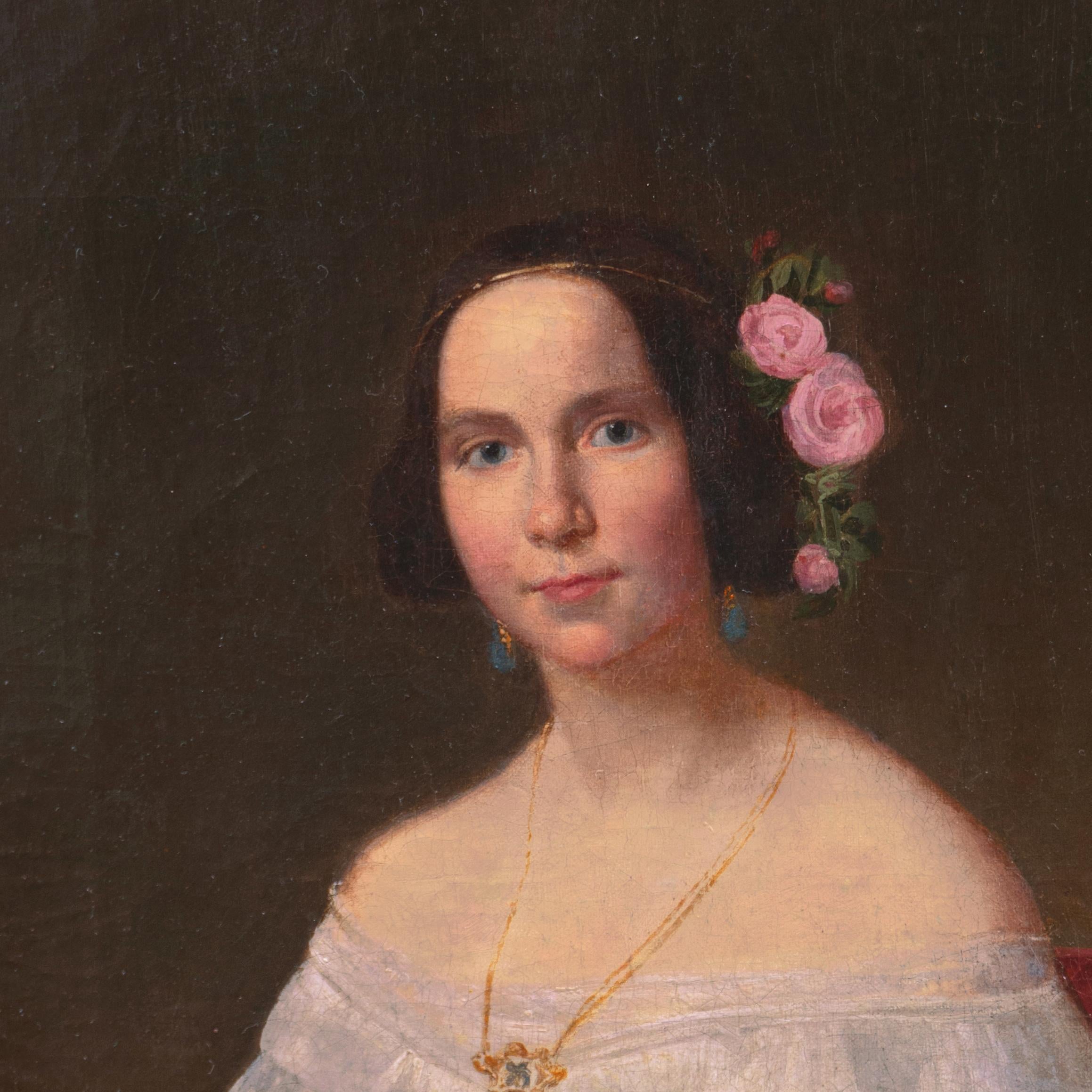 A delicately-observed, three-quarter length oil portrait of a young woman in society, fashionably dressed in a pinch-waisted, embroidered chiffon ball-gown and wearing a gold pendant and teardrop earrings with tea roses in her hair; the subject