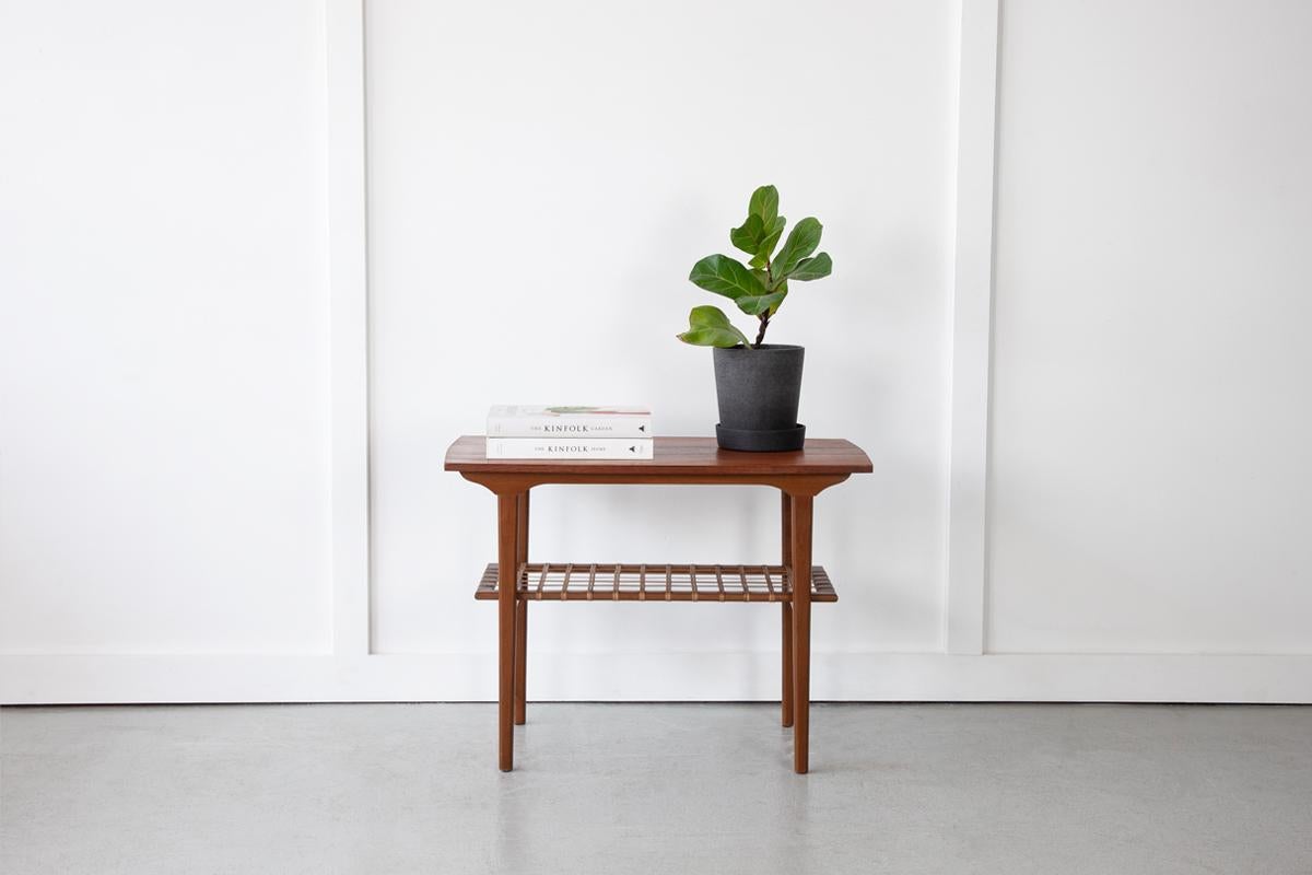 A neat Danish side table in teak with wonderful grain detailing and original woven cane magazine shelf. A stand-out piece for use as either as a living room side table or bedside table. 

Dimensions: Height 55cm / Width 75cm / Depth 42.5cm 