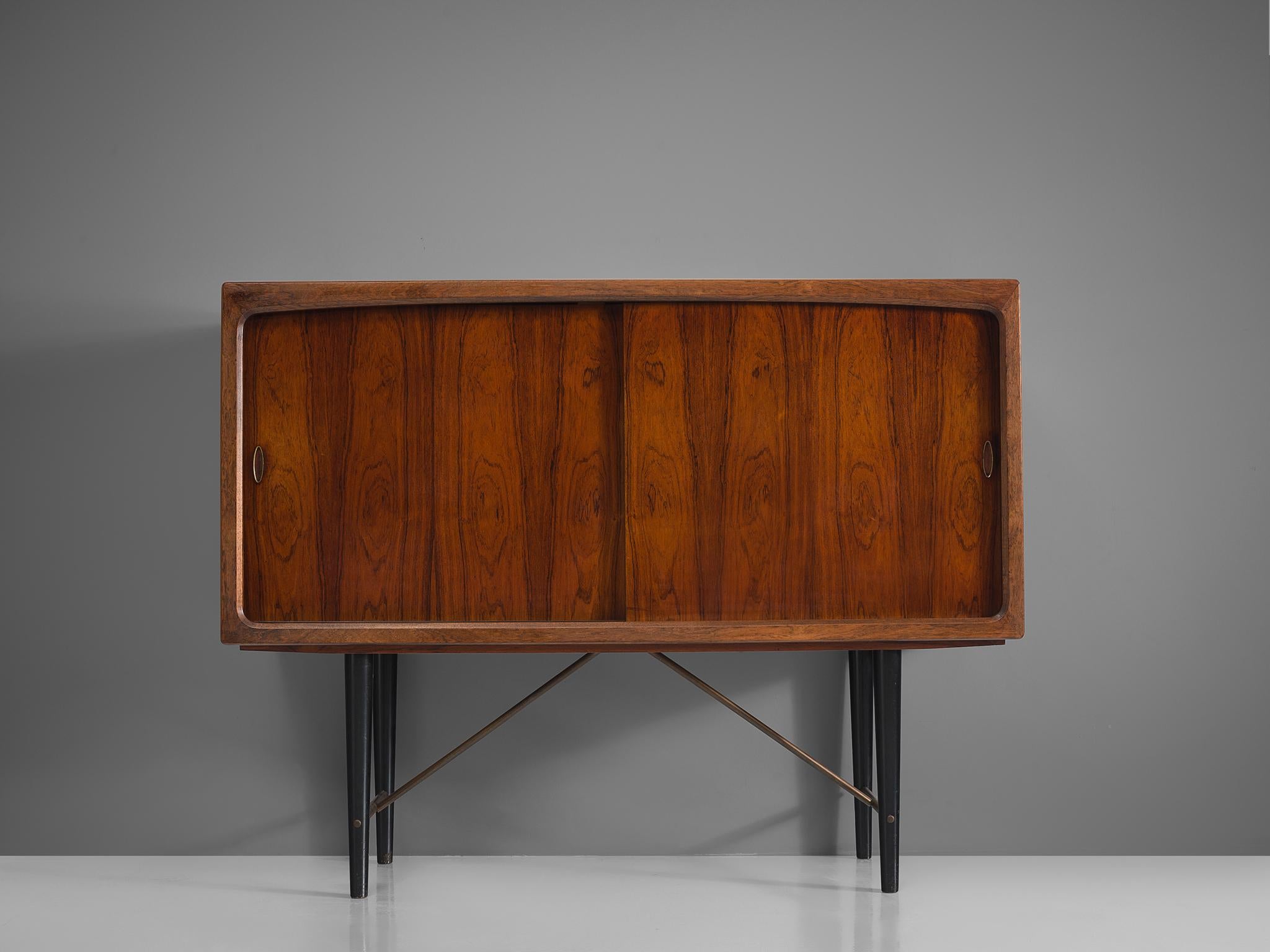 Danish sideboard, rosewood, metal, Denmark, 1950s

This cabinet is executed in rosewood and is typical for midcentury Scandinavian design. It has a base with four black wooden tapered legs that are connected via horizontal rods and brass diagonal