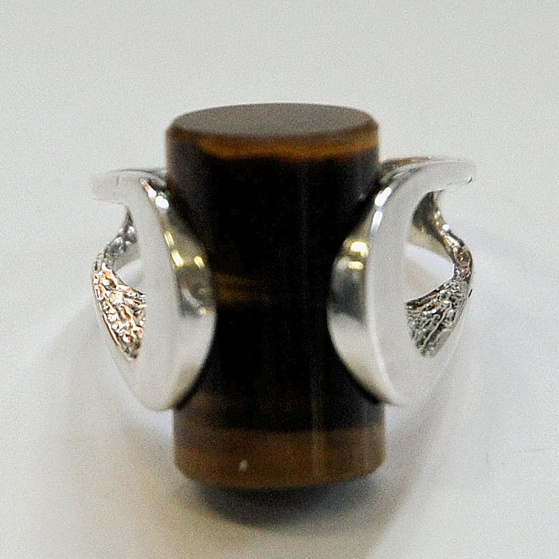 A fantastic shaped silver ring with a brown and black cylinder tiger eye stone embraced by an oval silver band holding the stone. Designed and made by Henning Ulrichsen, Fredericia in the 1970s Denmark.
Inner diameter is 18 mm, total height of ring