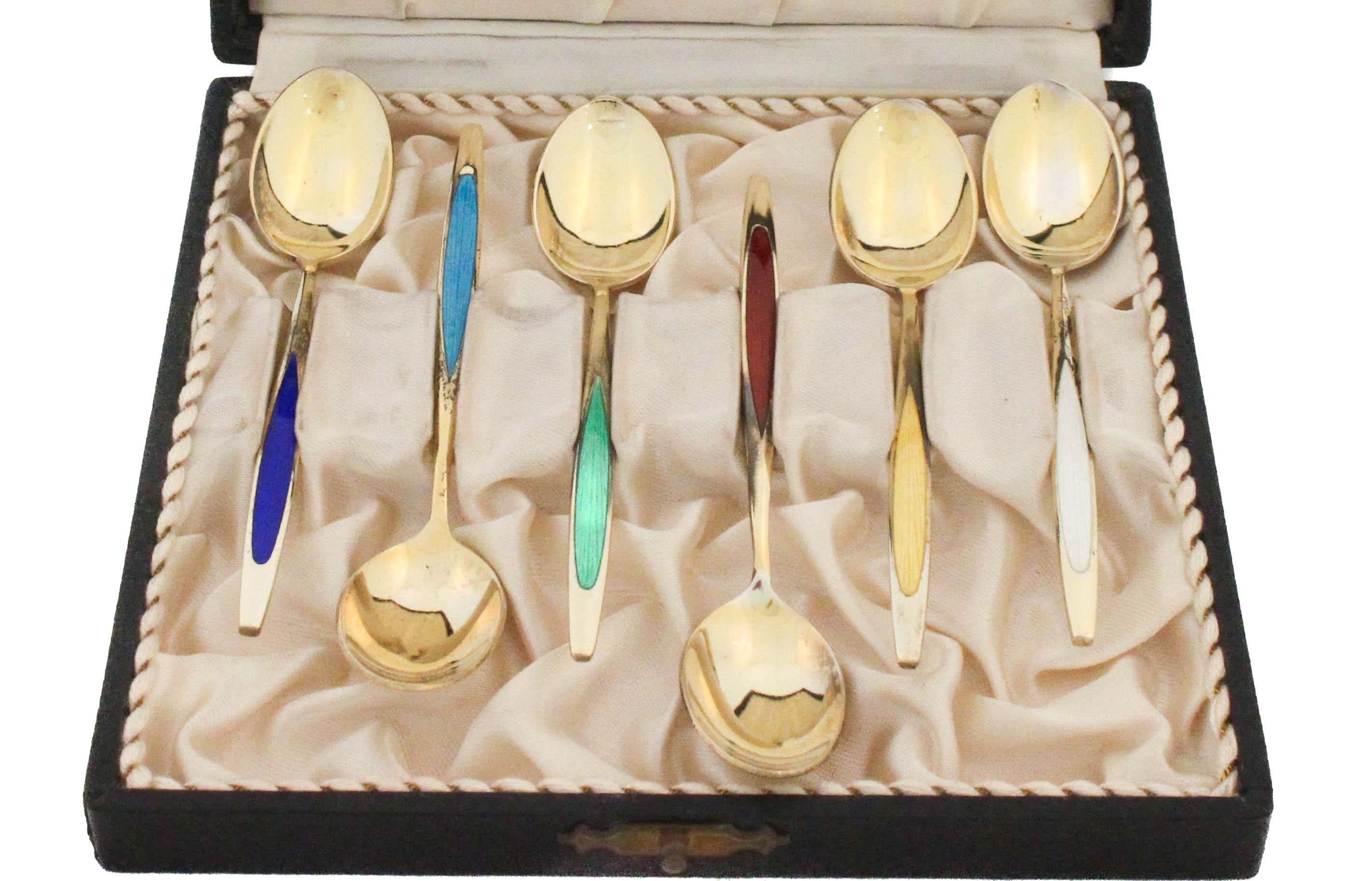 Il s'agit d'un ensemble de six cuillères à thé/démitasse en argent sterling et en émail, fabriquées au Danemark, présentées dans leur coffret d'origine. Chaque cuillère présente un design épuré et moderniste avec une finition dorée et un accent