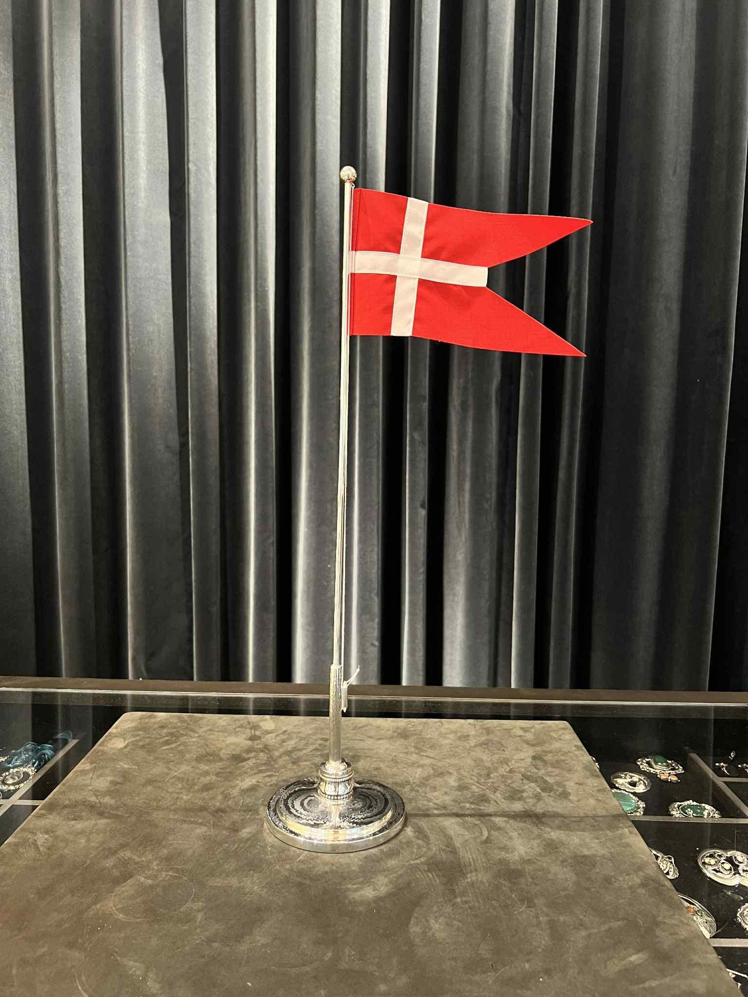 Una alta asta de bandera danesa de plata de ley, meticulosamente forjada para personificar los máximos niveles de calidad. Esta asta de bandera tiene una base redonda, martillada y escalonada que sirve de base robusta para el mástil. El mástil tiene
