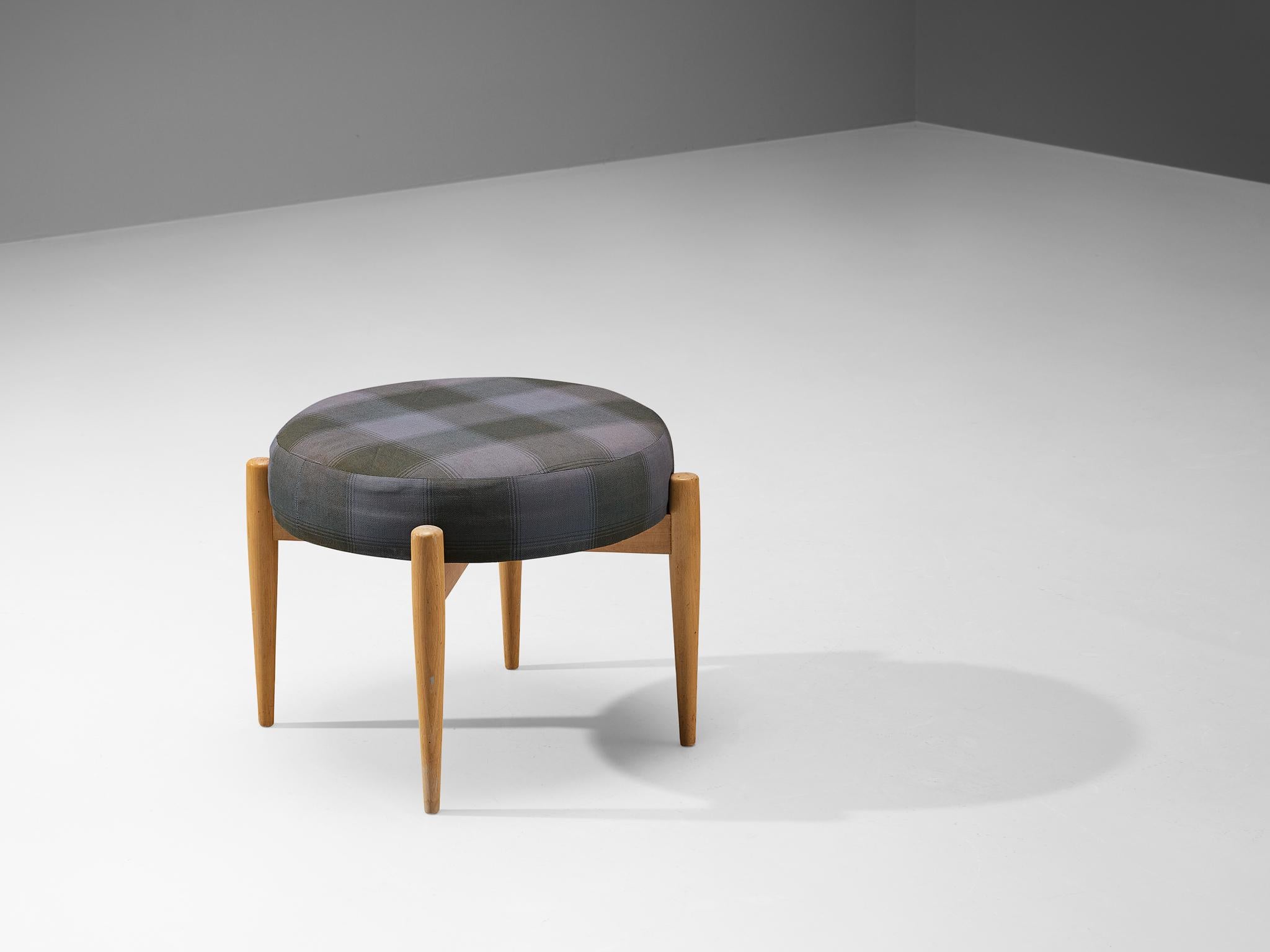 Stool or ottoman, beech, fabric, Denmark, 1960s

A Danish low stool with a round seat upholstered in a checkered fabric in black and grey hues. The base is composed of four round legs connected to a cross-shaped design. This well-proportioned piece