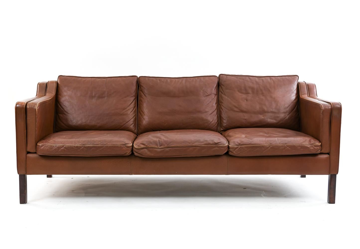 This Danish midcentury sofa was manufactured by Stouby. The design is in the manner of the iconic Borge Mogensen. This sofa is upholstered in attractively patinated brick colored leather and seats three comfortably.