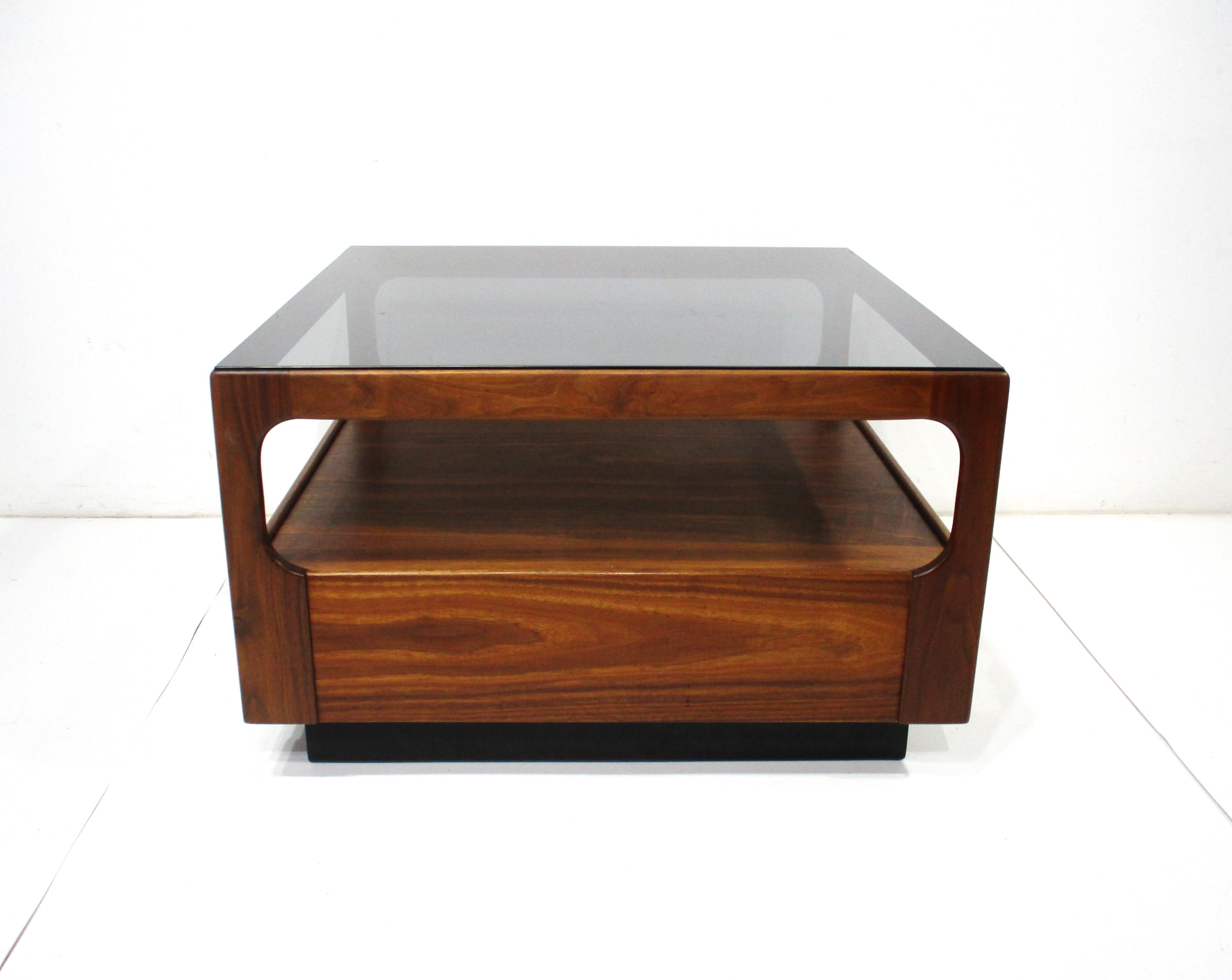 A medium dark walnut framed square coffee table with smoked glass inset top . The table has a lower shelving area for storage which makes it very useful for your living space . Crafted by Otmar in the manner of Danish Modern design with attention