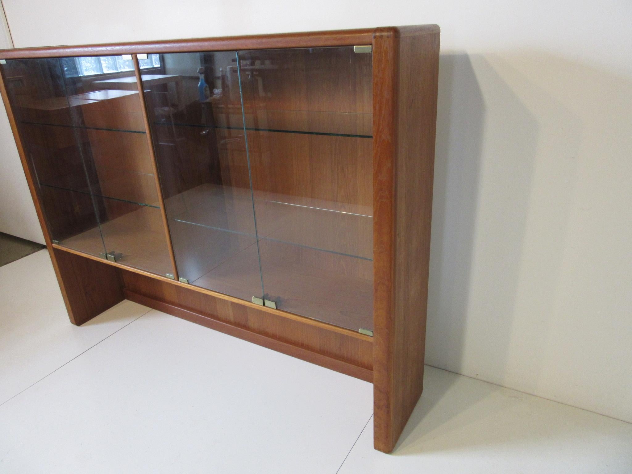 A medium toned teak wood bookcase with push touch glass doors and adjustable glass shelves. The glass doors have brass hardware and finger joint construction to the top corner pieces of the case manufactured by D-Scan imported from Singapore.