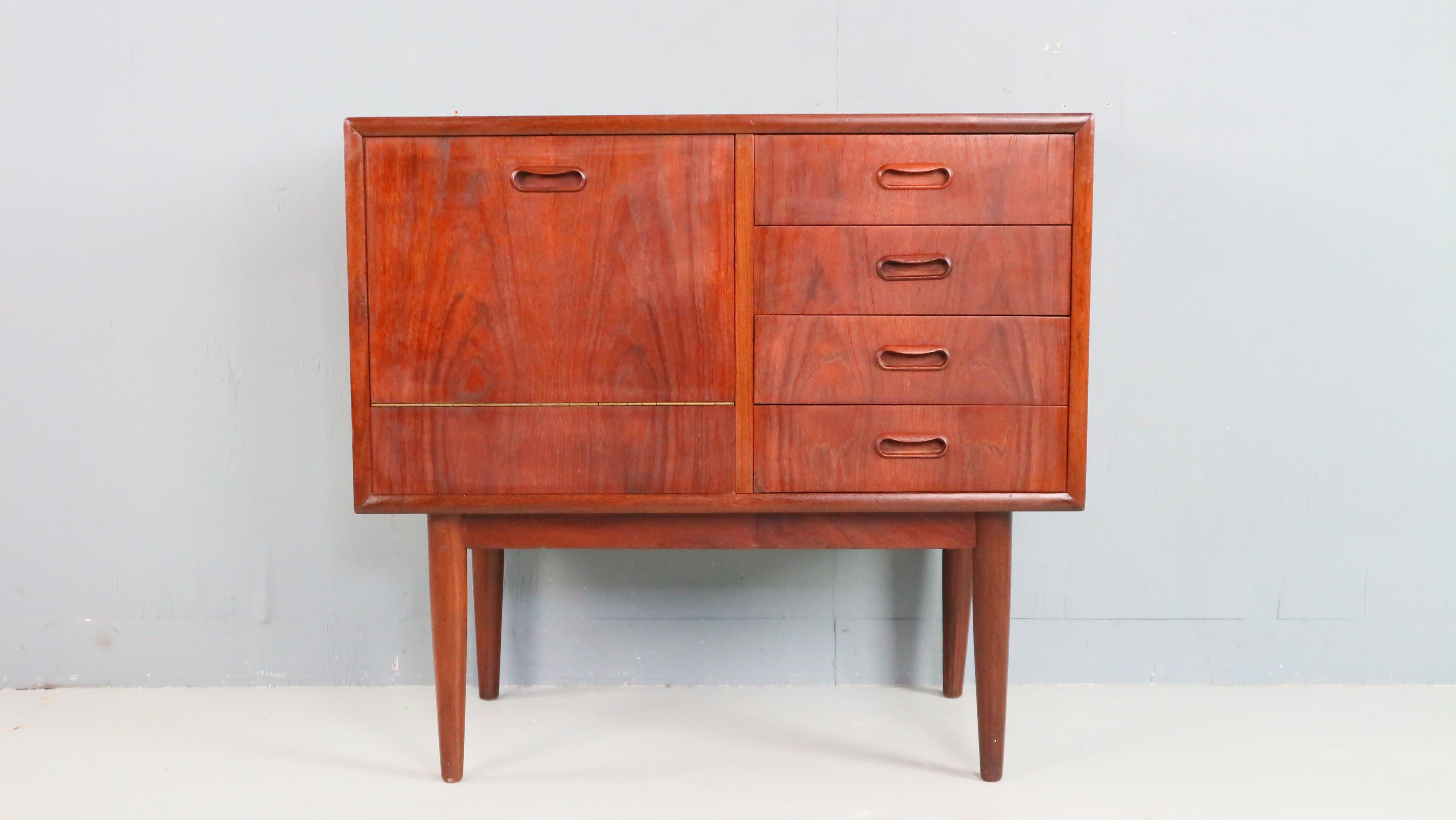 Charming Danish cabinet from the 1960s, made of warm teak. On the right, it features four small drawers; on the left, a drop-down door reveals additional storage space. The clean lines and carefully crafted wooden handles reflect the timeless